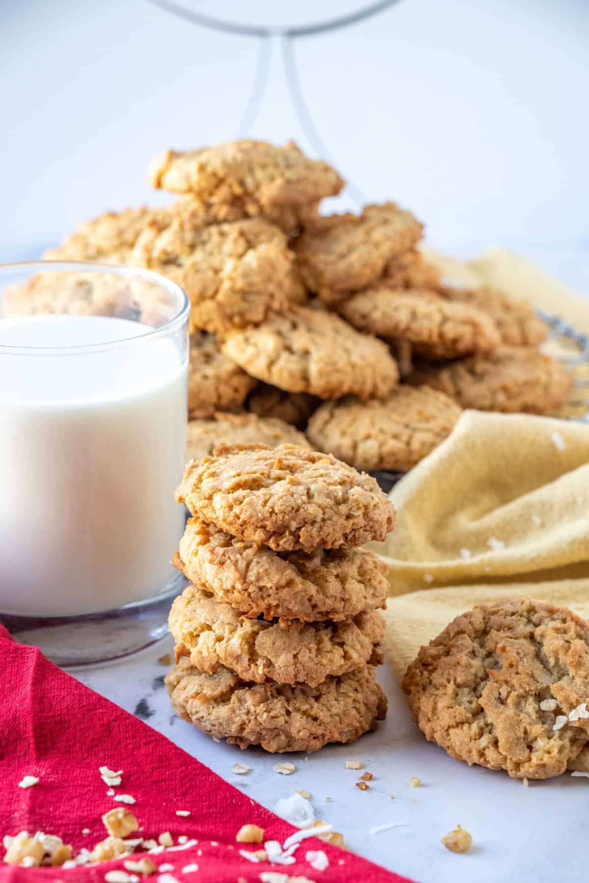 Four stacked cookies with a glass of milk and a platter of cookies in the background. 