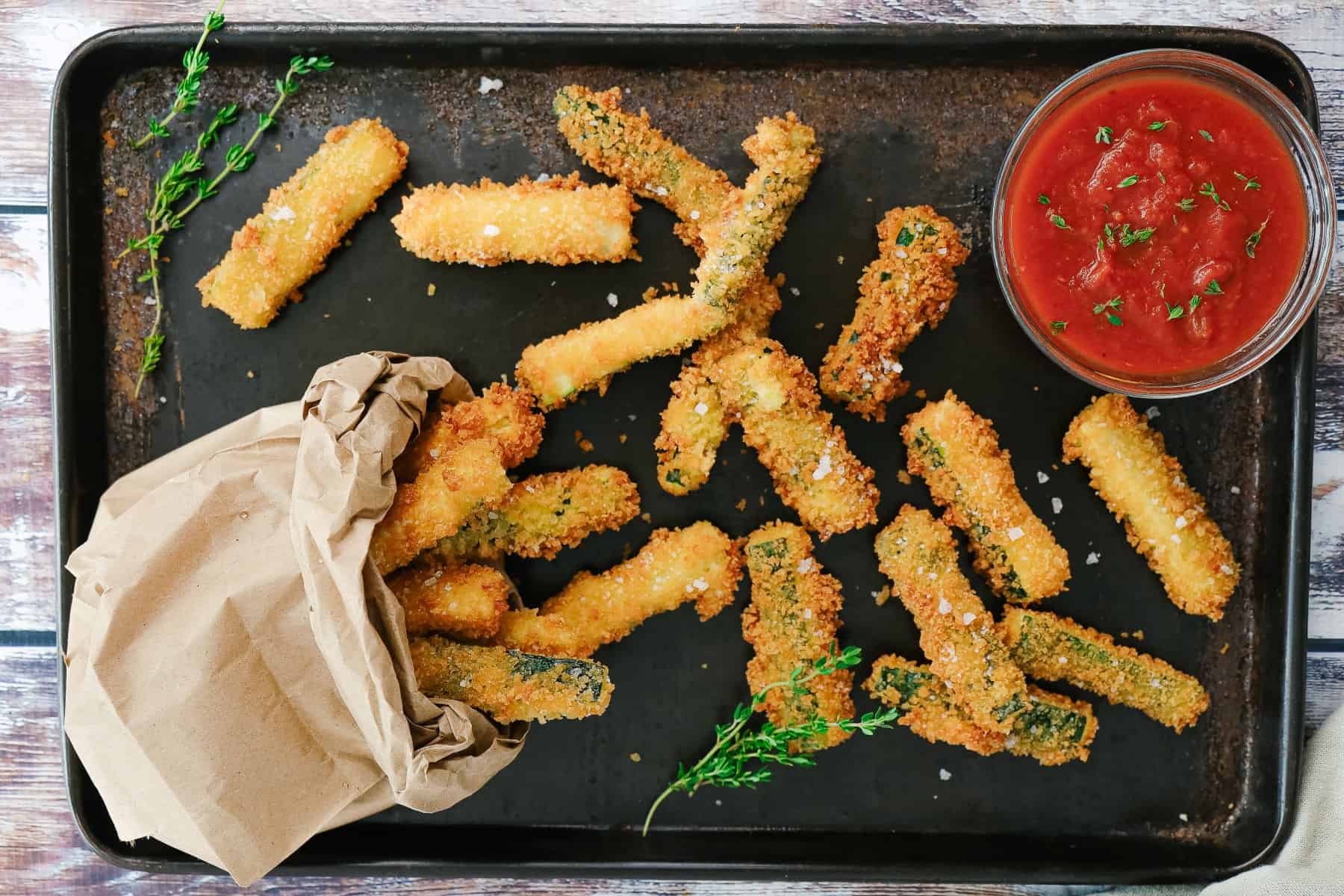 Zucchini sticks on a baking tray and in a paper bag with marinara sauce. 