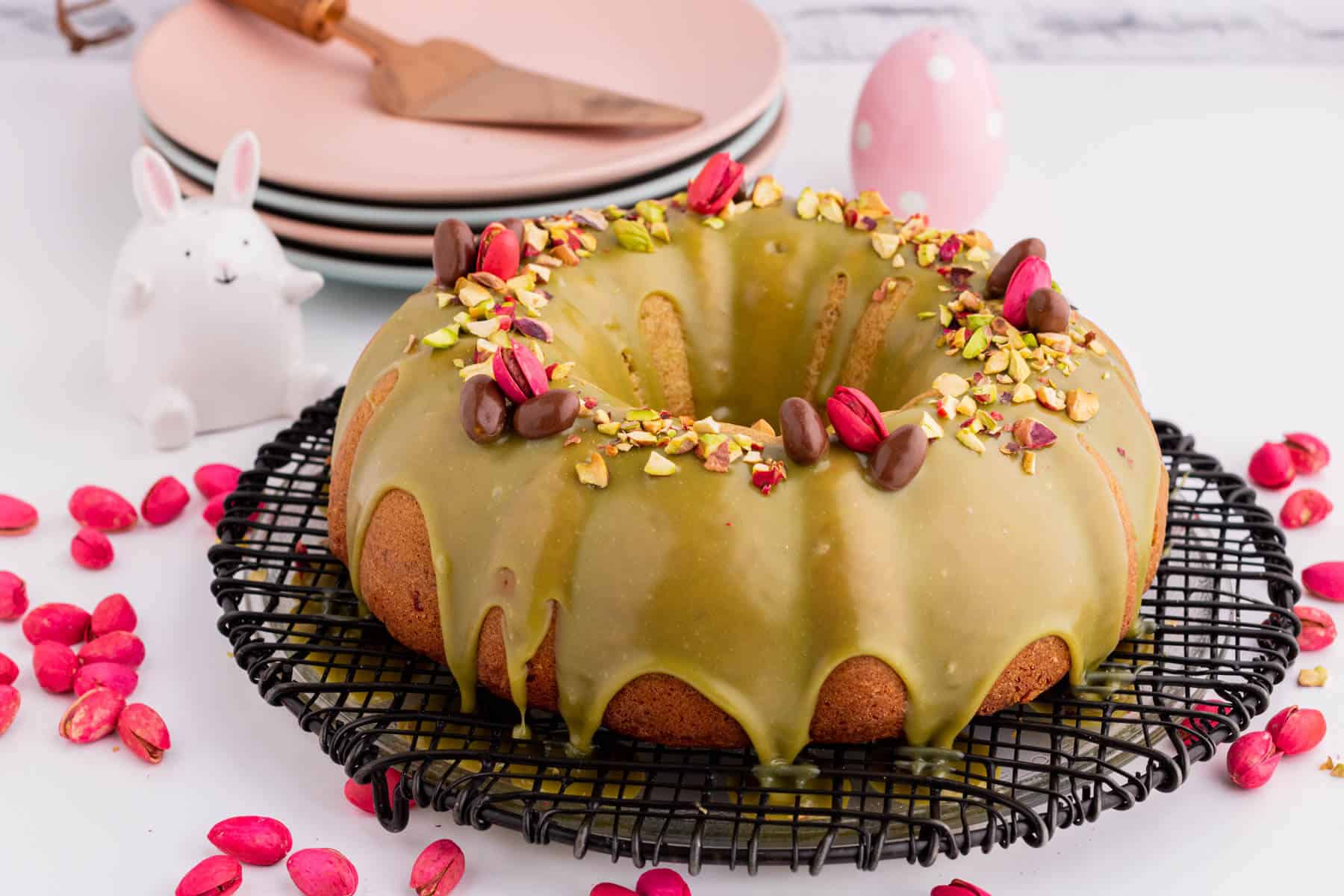 A green glazed bundt cake decorated with chopped pistachios, whole pink and chocolate pistachios.