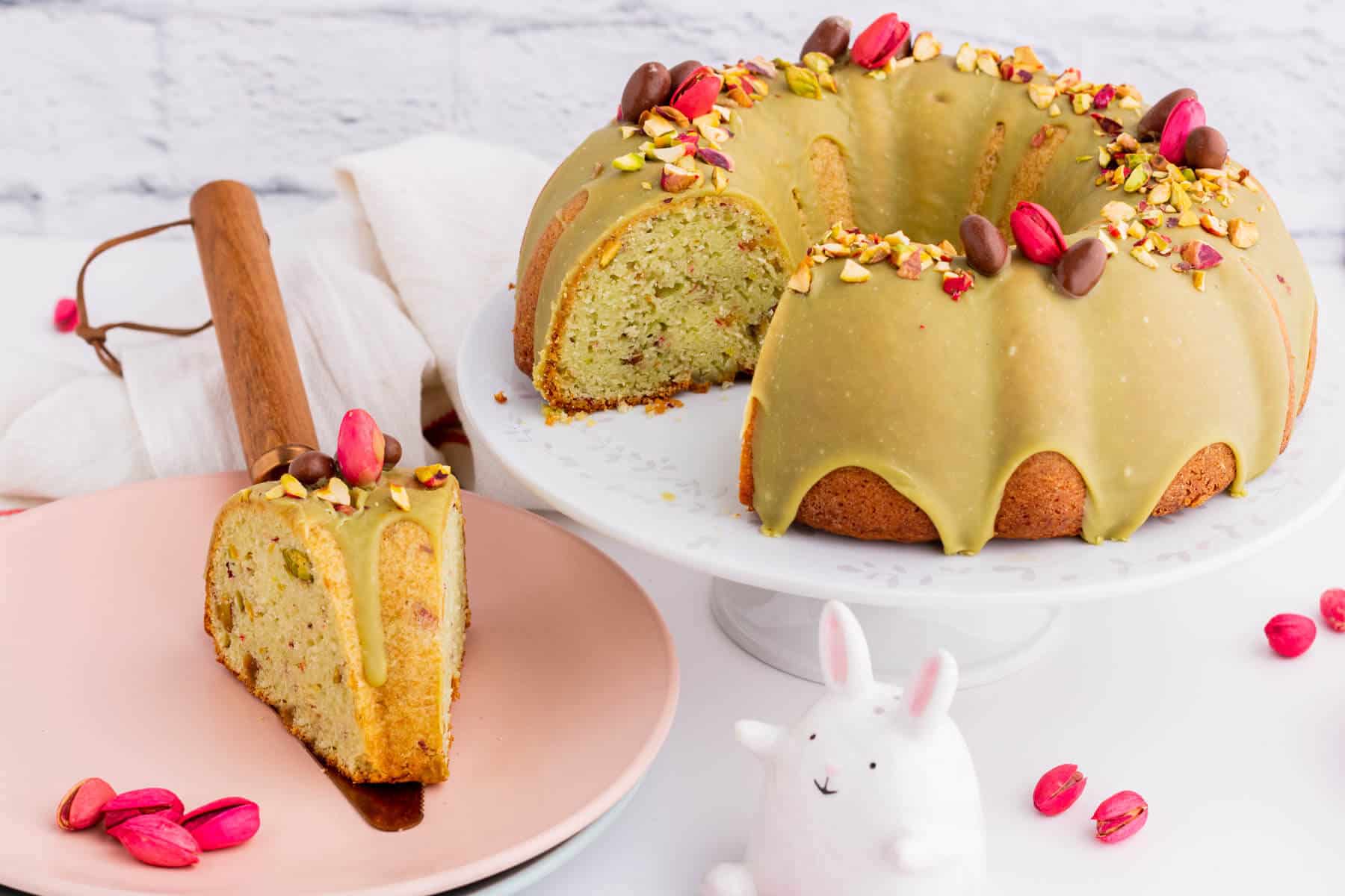 A slice of cake on a plate beside a whole pistachio decorated bundt cake. 