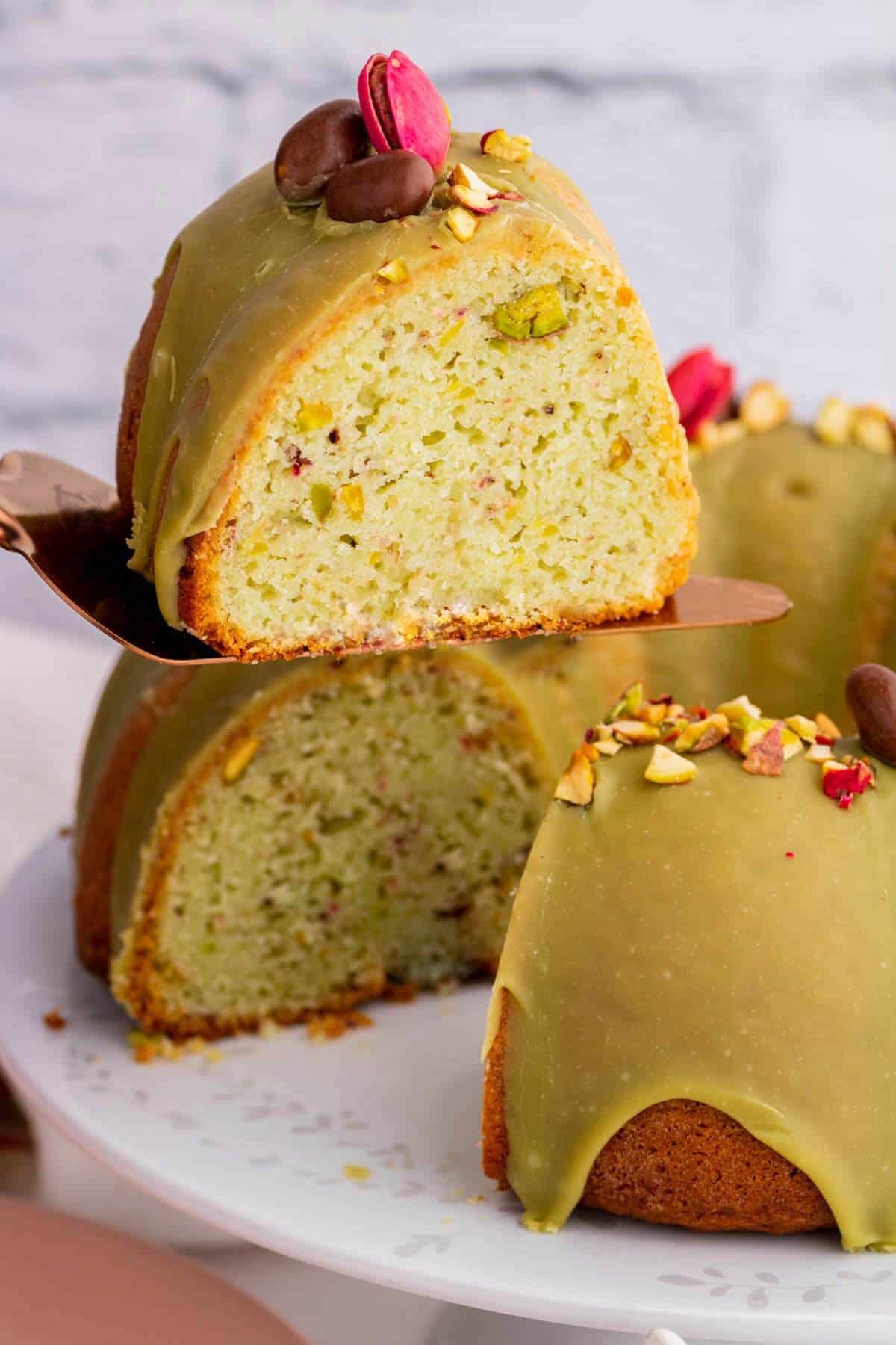 A slice being served from a green glazed bundt cake decorated with pink and chocolate pistachios.
