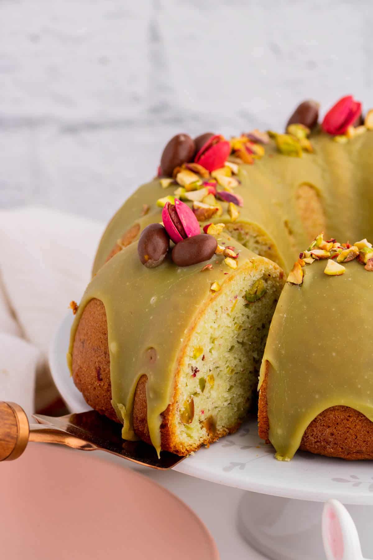 A slice being served from a green glazed bundt cake decorated with pink and chocolate pistachios.