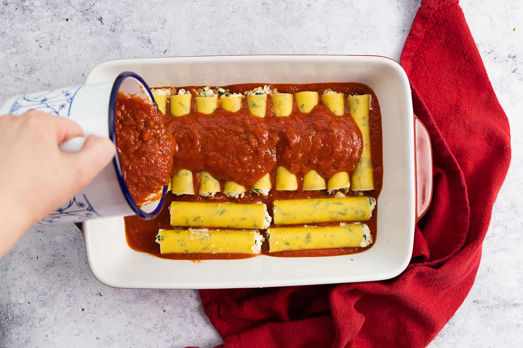 Top view of a baking dish filled with uncooked manicotti and a hand pouring marinara sauce.