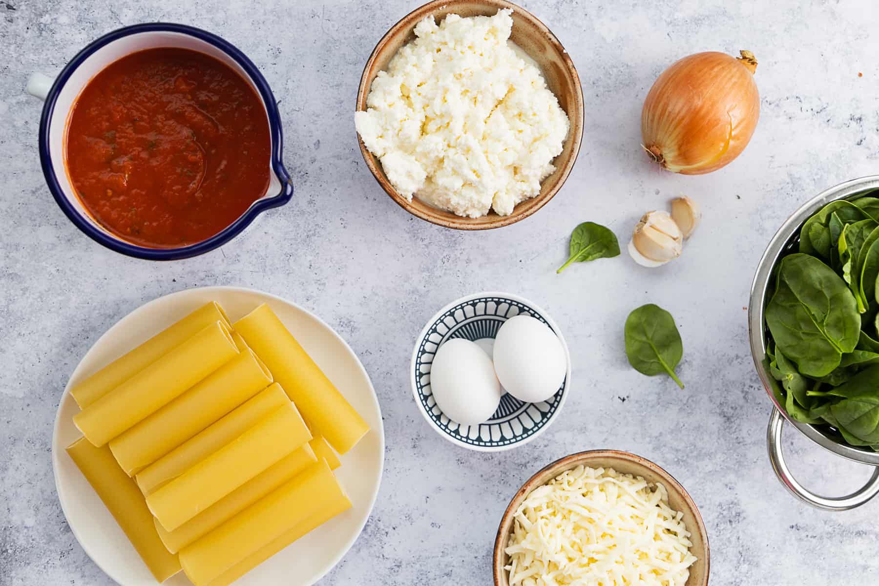 Top view of ingredients for cheese manicotti: pasta shells, tomato sauce, cheeses, eggs, onion, garlic and spinach. 