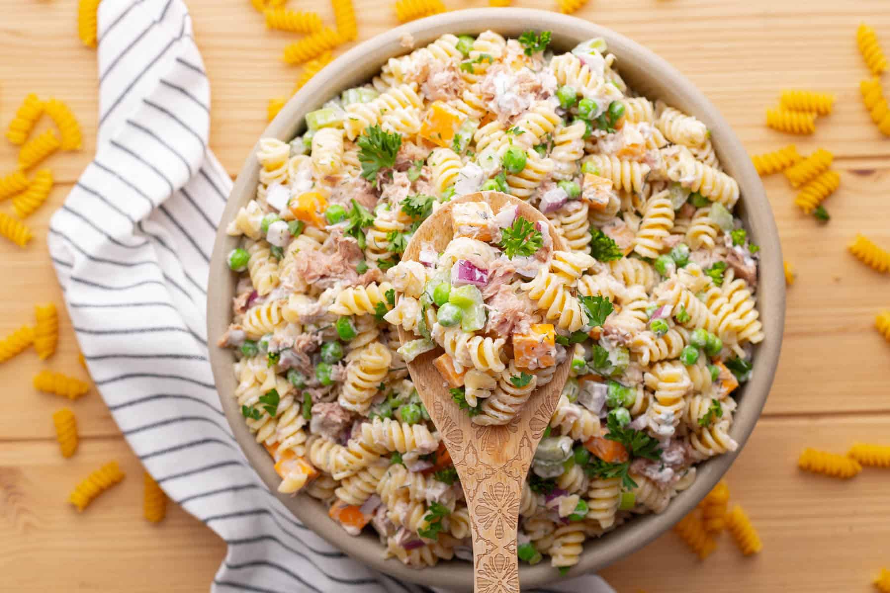 A wooden spoon holds tuna pasta salad above a bowl filled with the same salad.