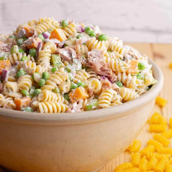 A serving bowl filled with rotini pasta, peas, tuna chunks, red onions, orange cheddar, and celery.