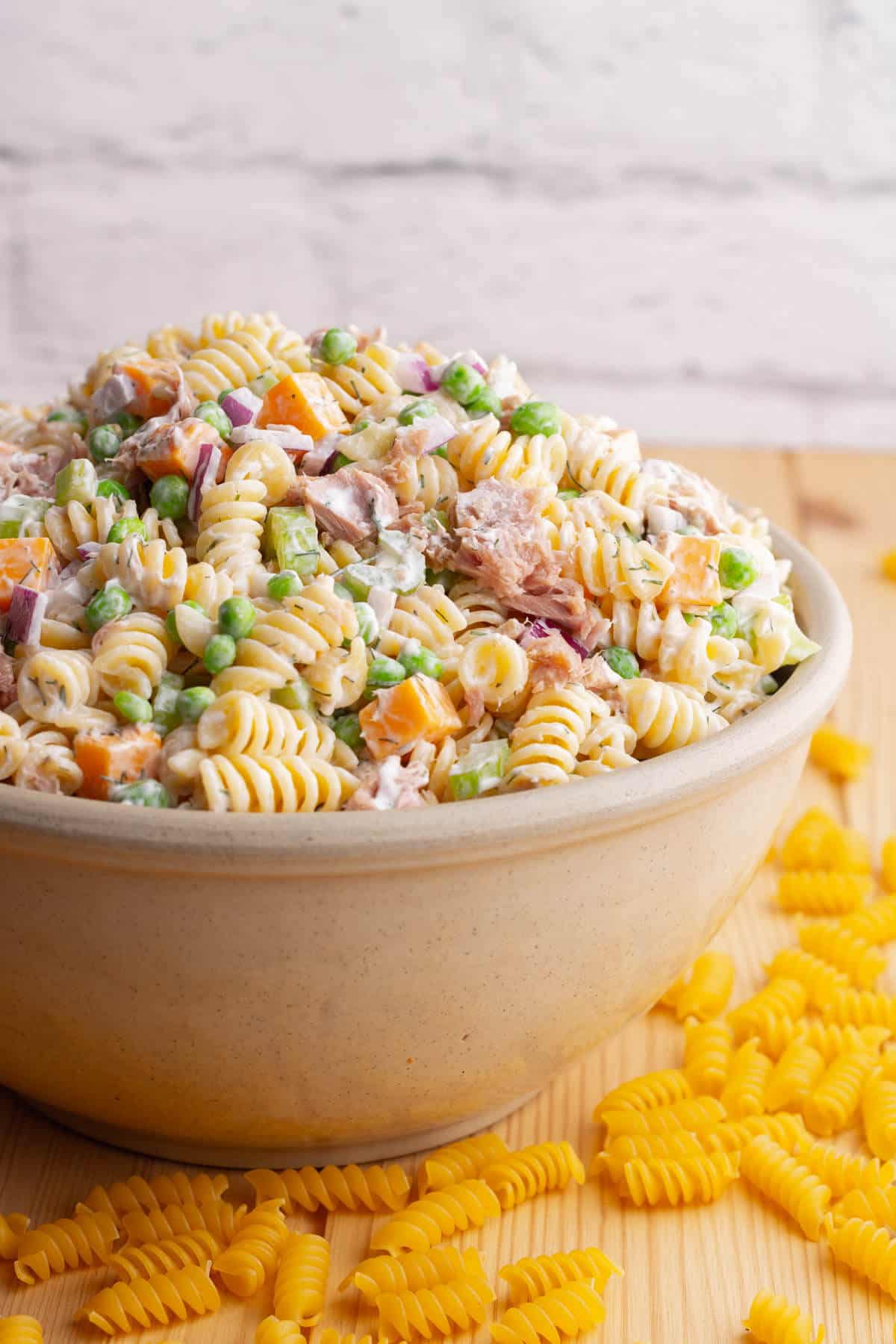 A serving bowl filled with rotini pasta, peas, tuna chunks, red onions, orange cheddar, and celery. 