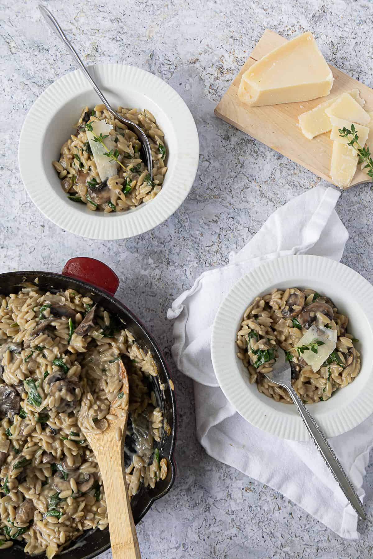 Top view of 2 bowls with mushroom orzo and a skillet on the side and a wood board with parmesan cheese on the other side. 