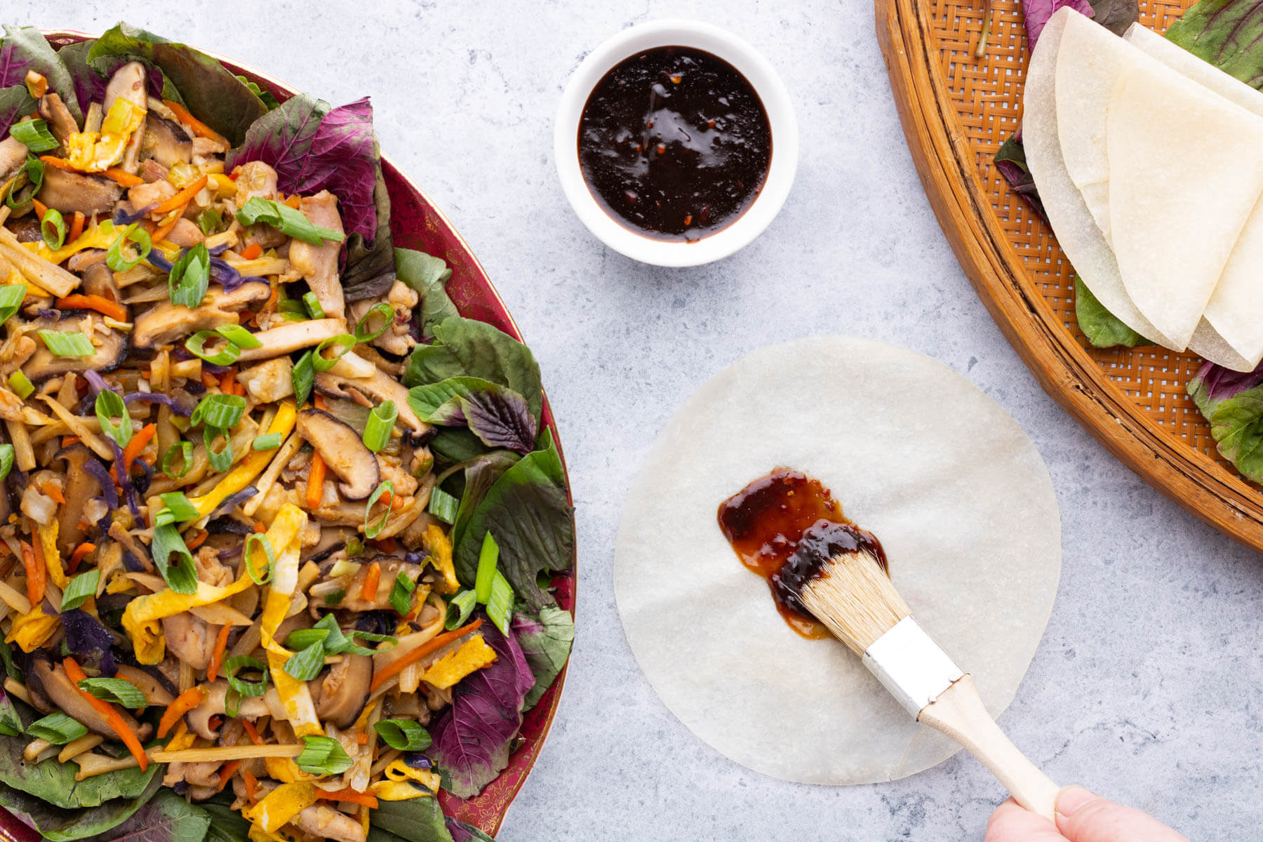 A brush paints hoisin sauce on a Chinese pancake beside a platter full of colourful Moo Shu Chicken.