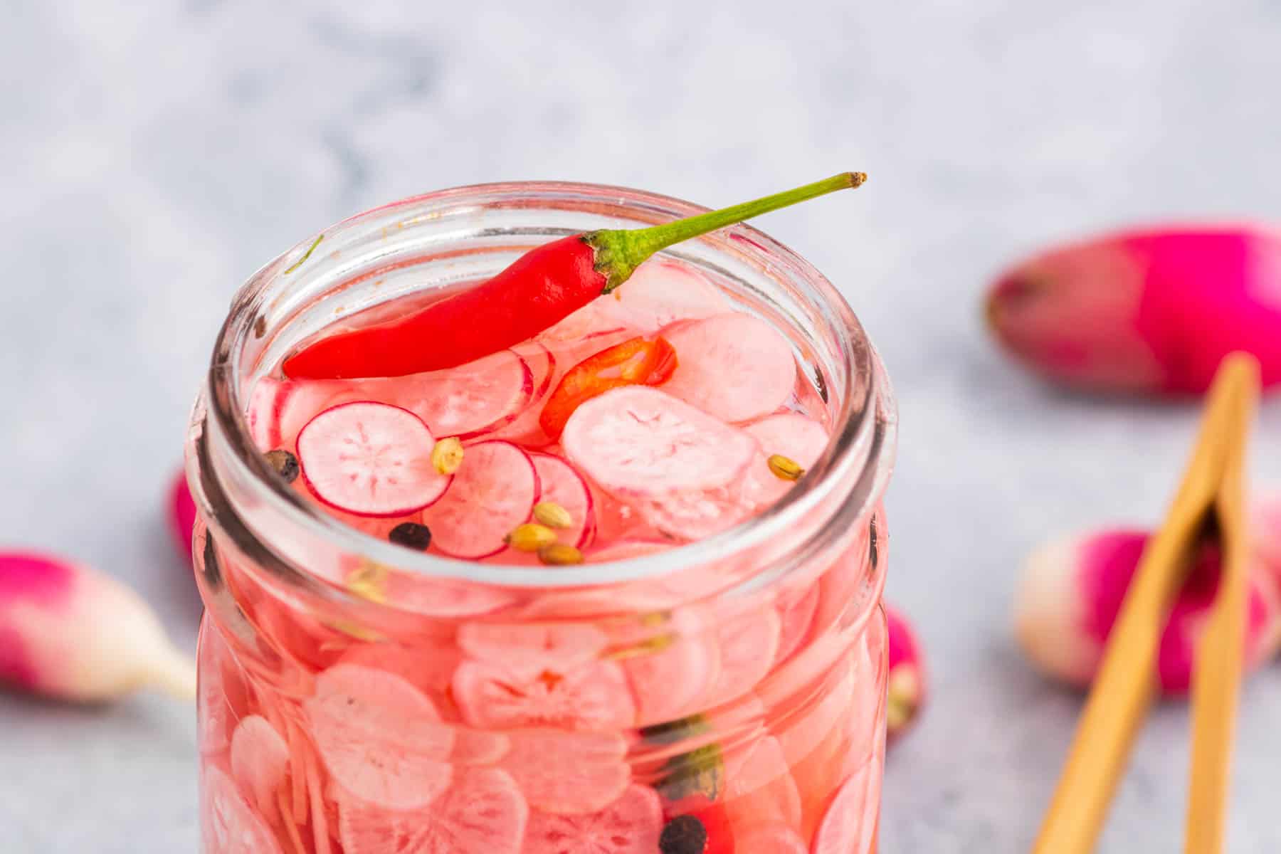The top of an open glass jar filled with sliced radishes, red chili pepper, peppercorns, and coriander seeds.