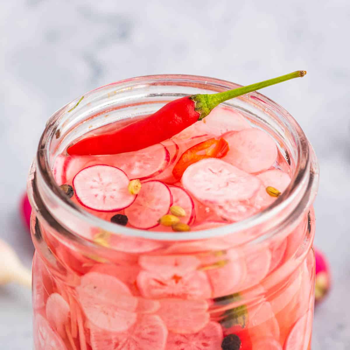 The top of an open glass jar filled with sliced radishes, red chili pepper, peppercorns, and coriander seeds.