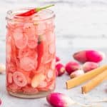 A half pint glass jar filled with sliced radishes, red chilis, peppercorns, coriander, and a garlic clove.