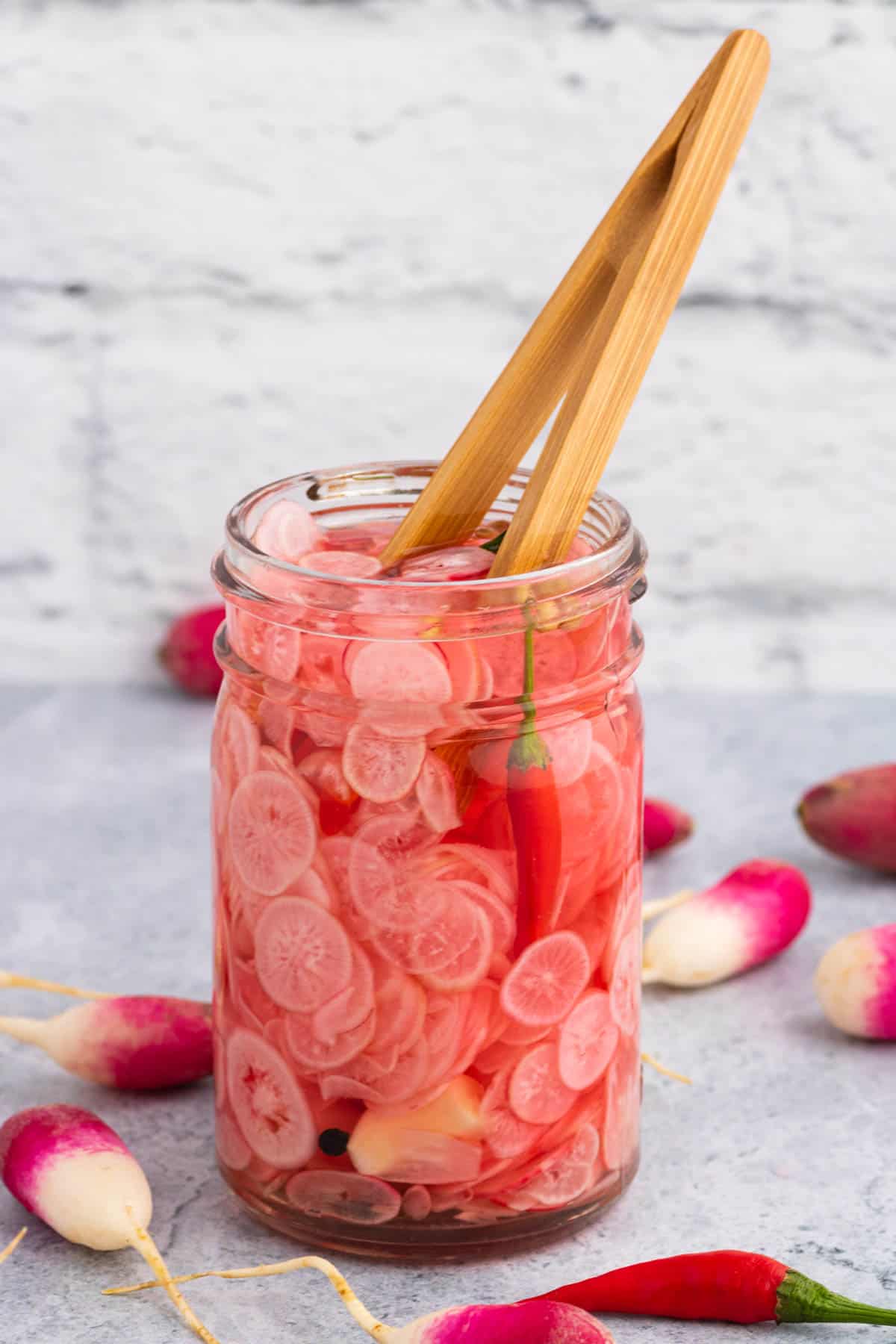 A half-pint glass jar filled with quick pickled radishes and a pair of wooden tongs.
