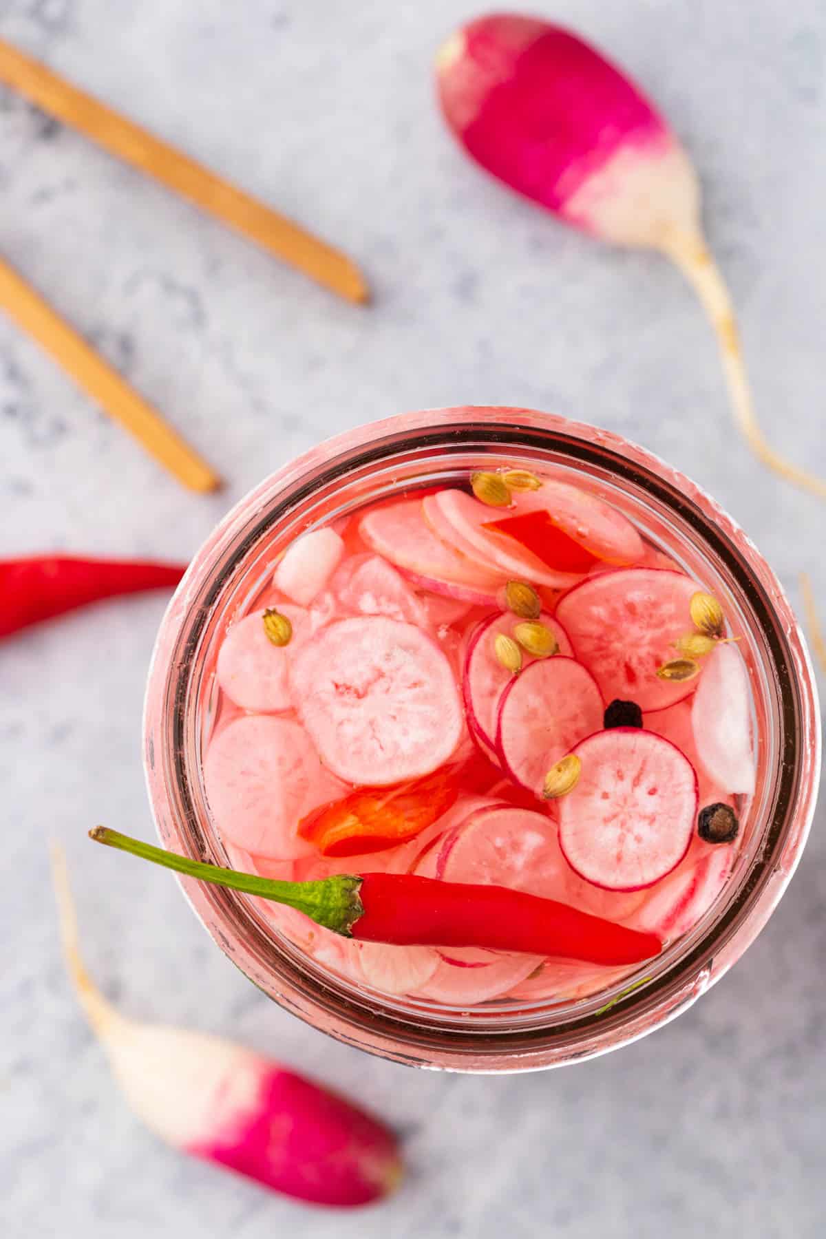 The top of an open glass jar filled with sliced radishes, red chili pepper, peppercorns, and coriander seeds.