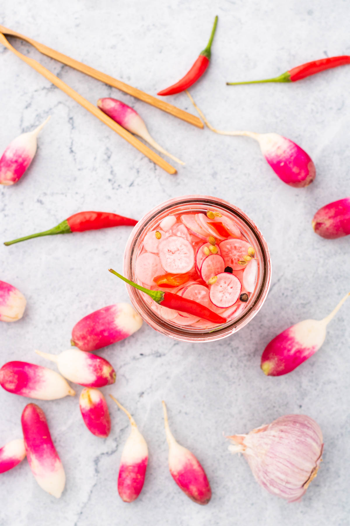 The top of an open glass jar filled with sliced radishes, red chili pepper, peppercorns, and coriander seeds. Surrounded by bright radishes, garlic, and red chilis.