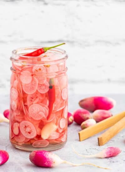 A half pint glass jar filled with sliced radishes, red chilis, peppercorns, coriander, and a garlic clove.