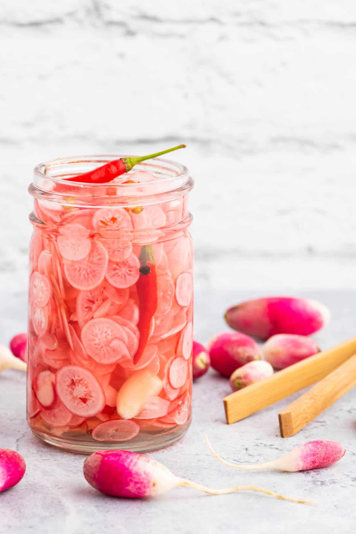 A half-pint glass jar filled with sliced radishes, red chilis, peppercorns, coriander, and a garlic clove.