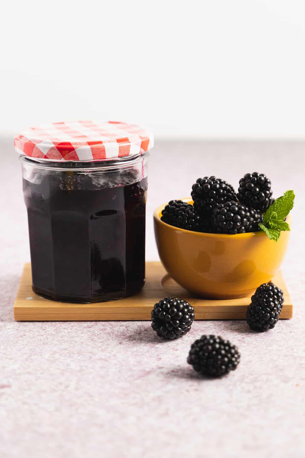 Side view of a glass jar of blackberry freezer jam closed and a yellow bowl with fresh berries on the side. 
