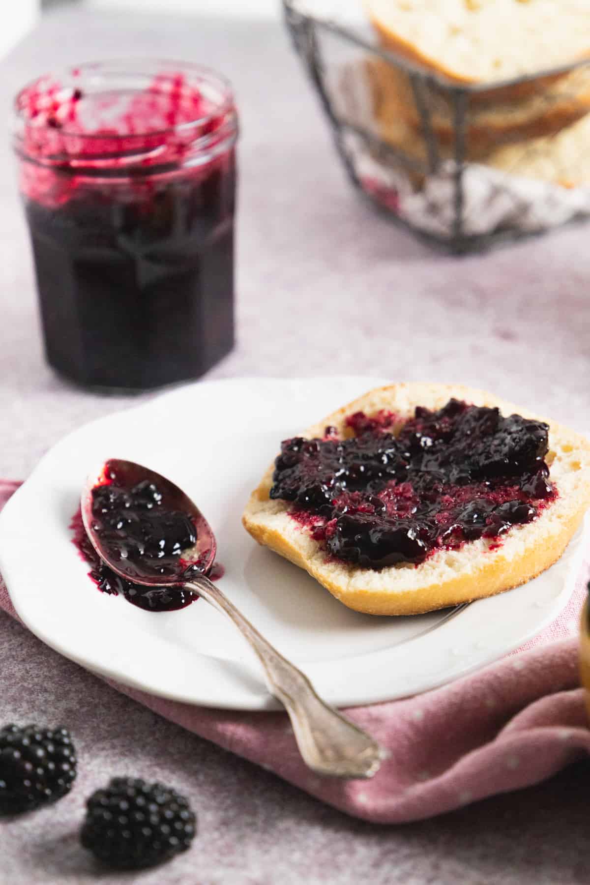 Side view of a white plate with a slice of bread covered with blackberry freezer jam and spoon on the side, a glass jar in the back.
