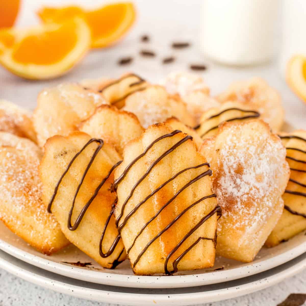 Plate of decorated (chocolate drizzle or icing sugar) Orange Madeleine Cookies. 