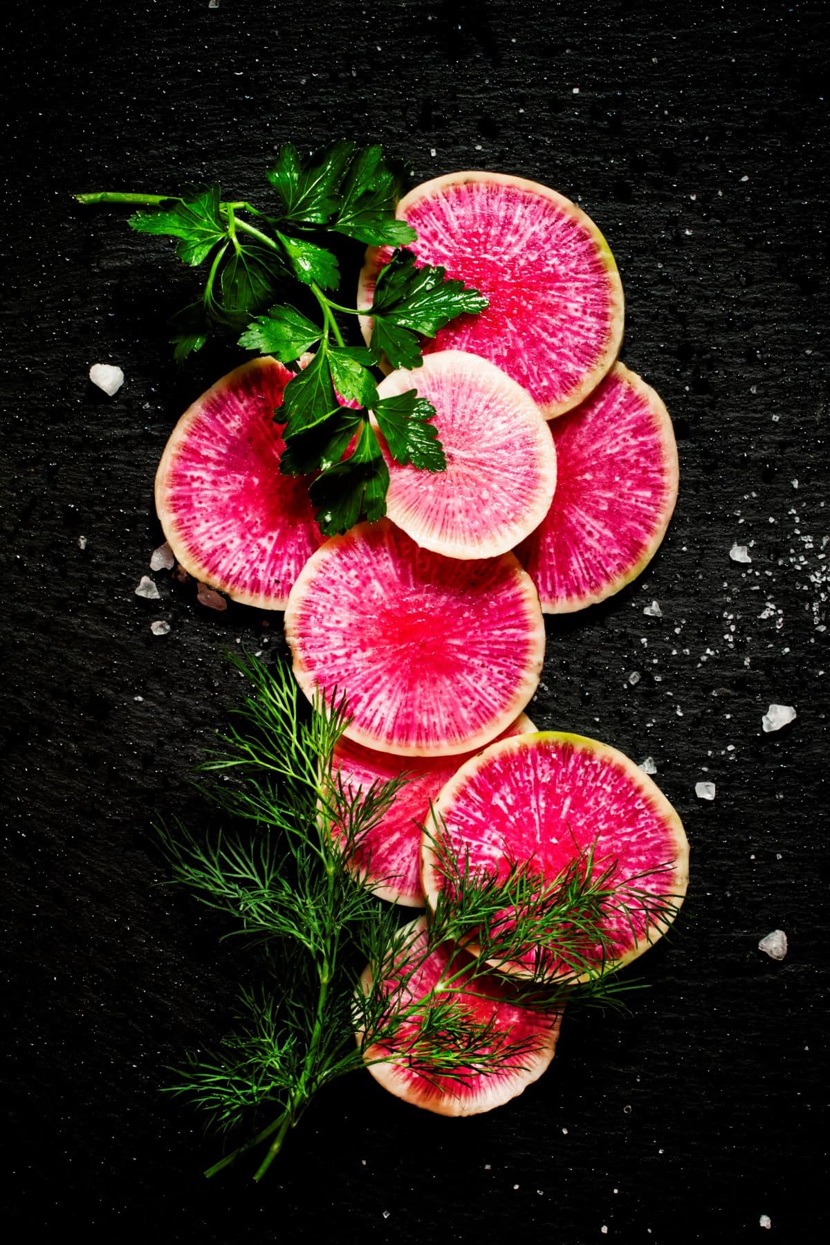 Food background: round slices of watermelon pink radish, dill, parsley, salt, black stone background, top view