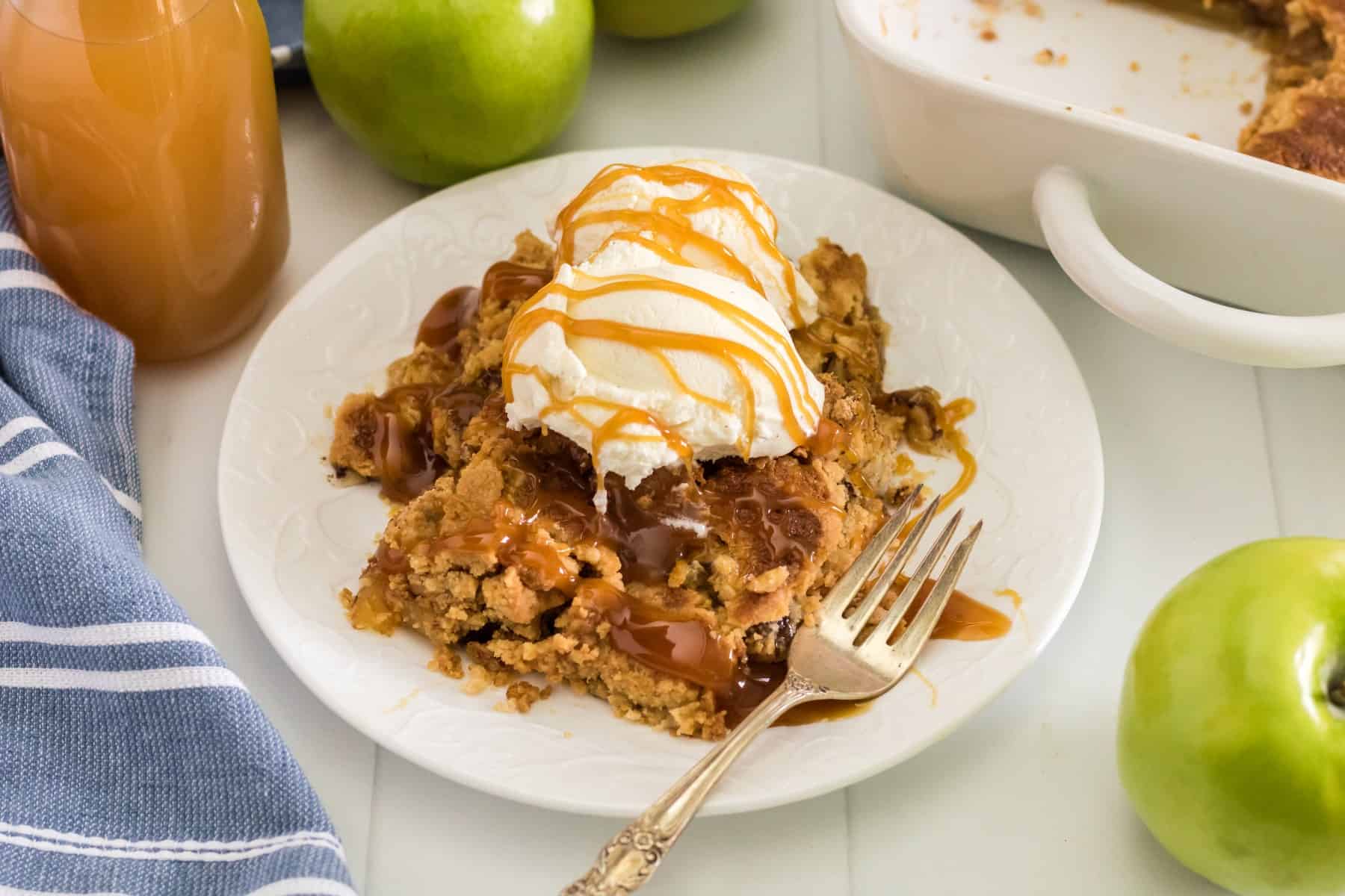 Horizontal photo of apple dump cake on a plate with ice cream and caramel sauce with apple cider in bottle and green apples around. 