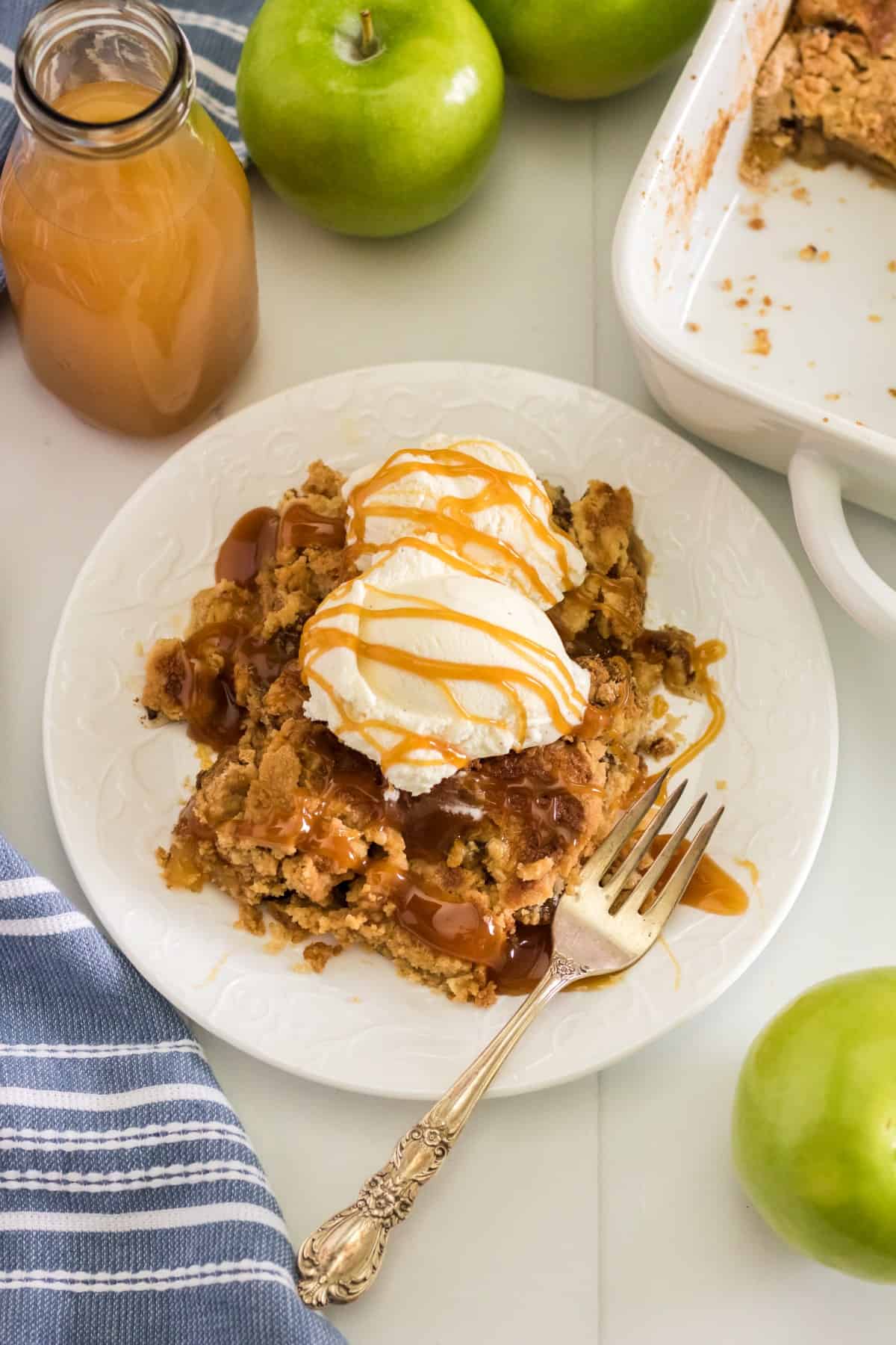 Slight overhead shot of apple dump cake on a plate with ice cream and caramel sauce with apple cider in bottle and green apples around. 