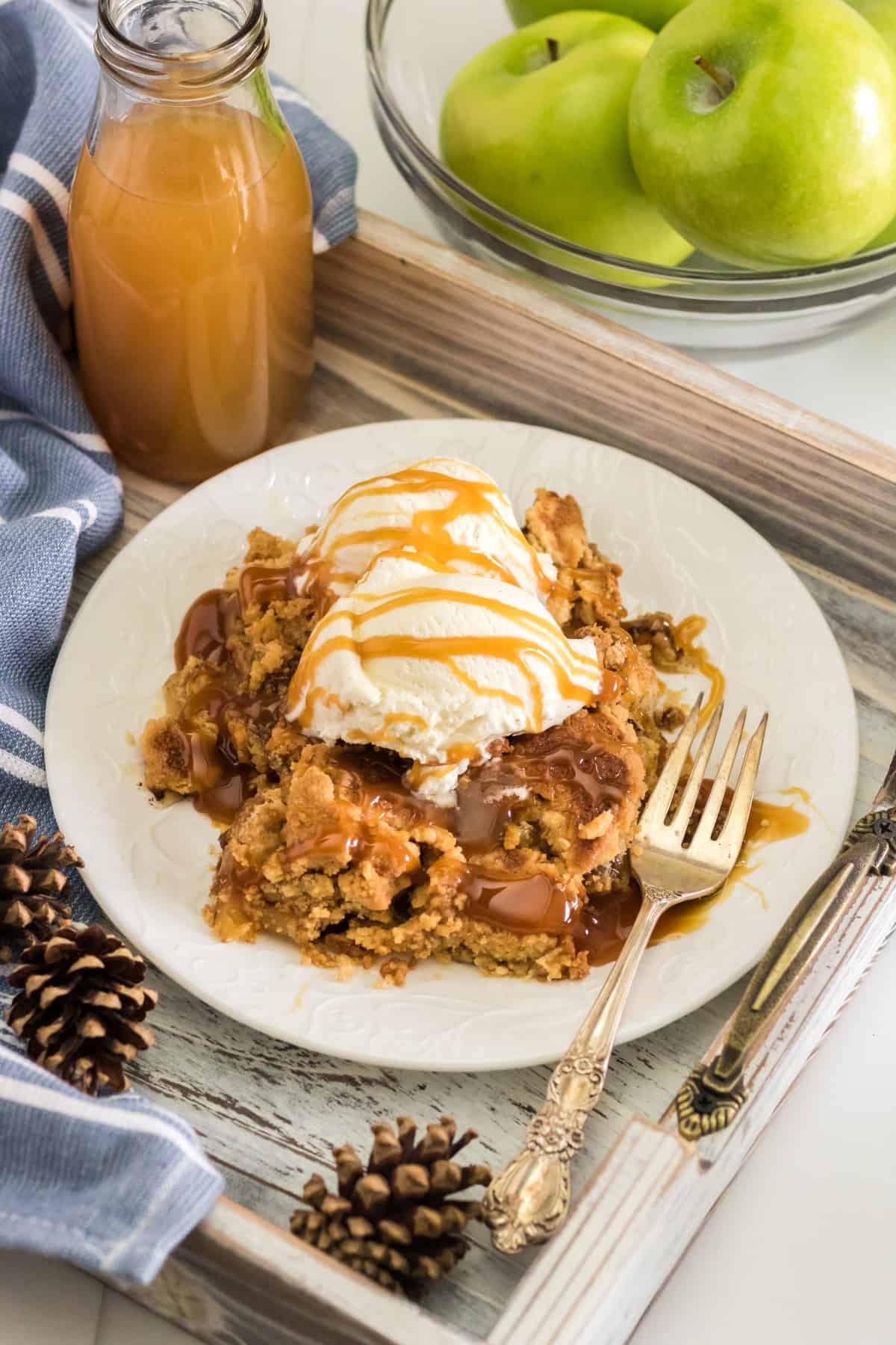 Apple dump cake with vanilla ice cream on a plate with caramel sauce on a plate that is on a tray with apple cider in a bottle and green apples in a bowl. 