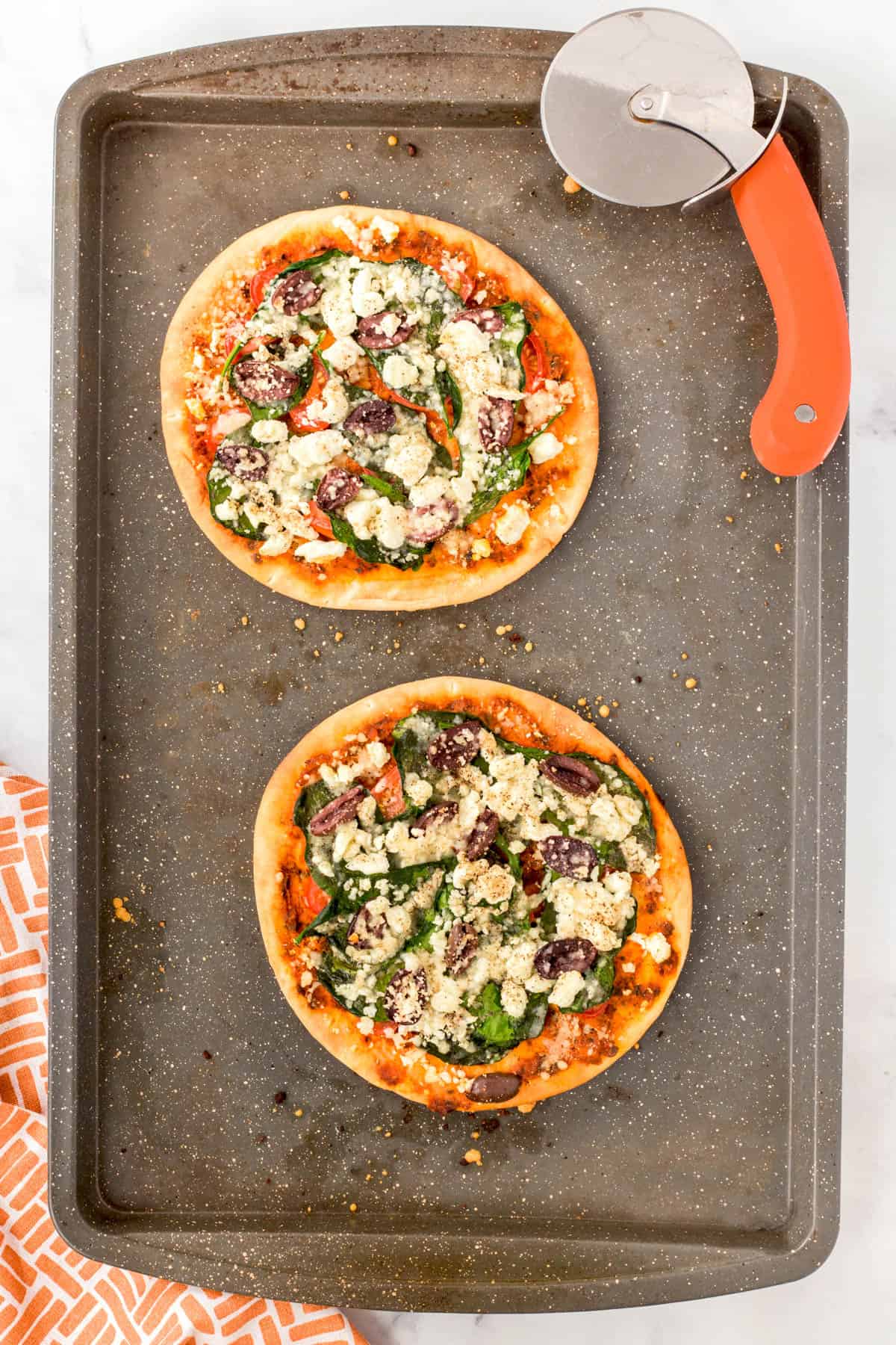 Two Greek Pita Pizzas on a baking sheet with a pizza cutter