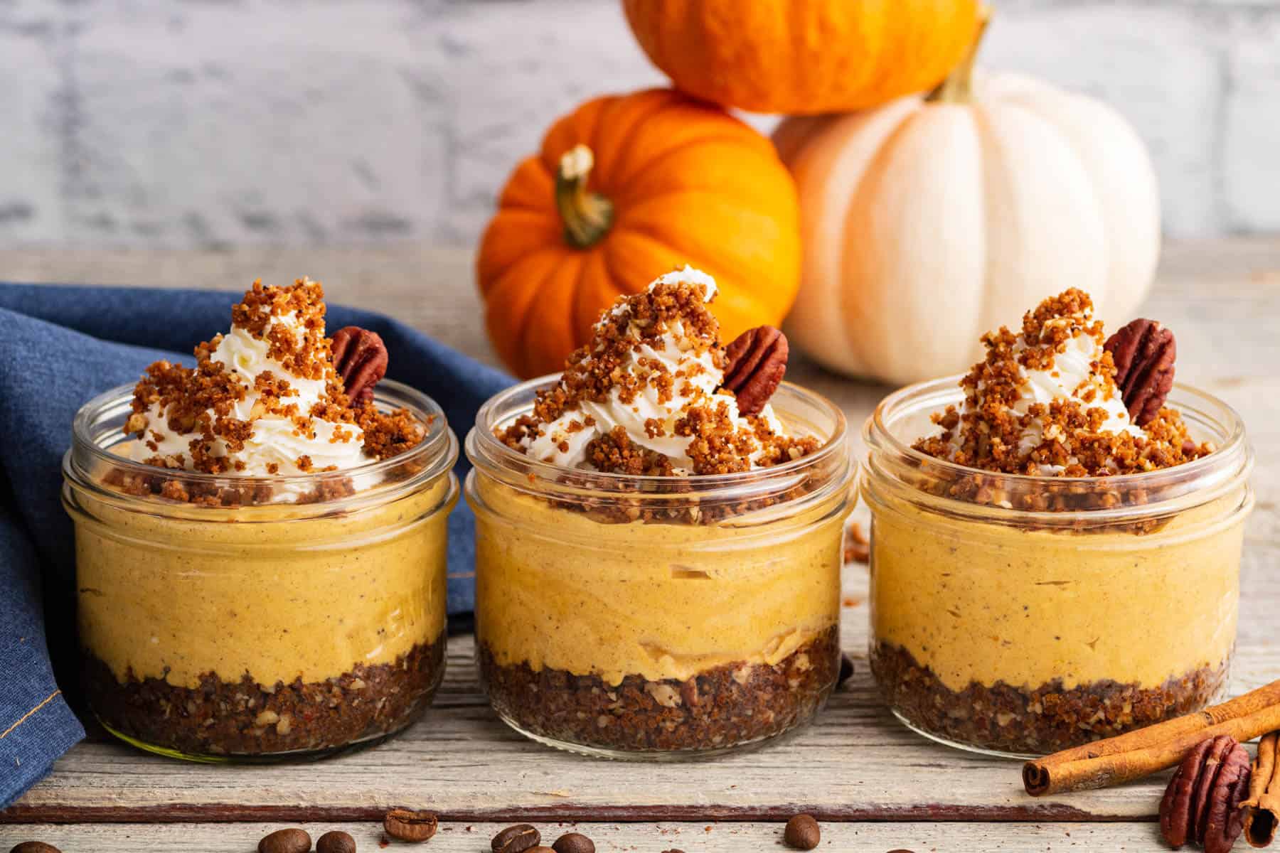 Three individually sized Pumpkin Delight desserts topped with gingersnap crumbs and pecans sit amongst decorative pumpkins.