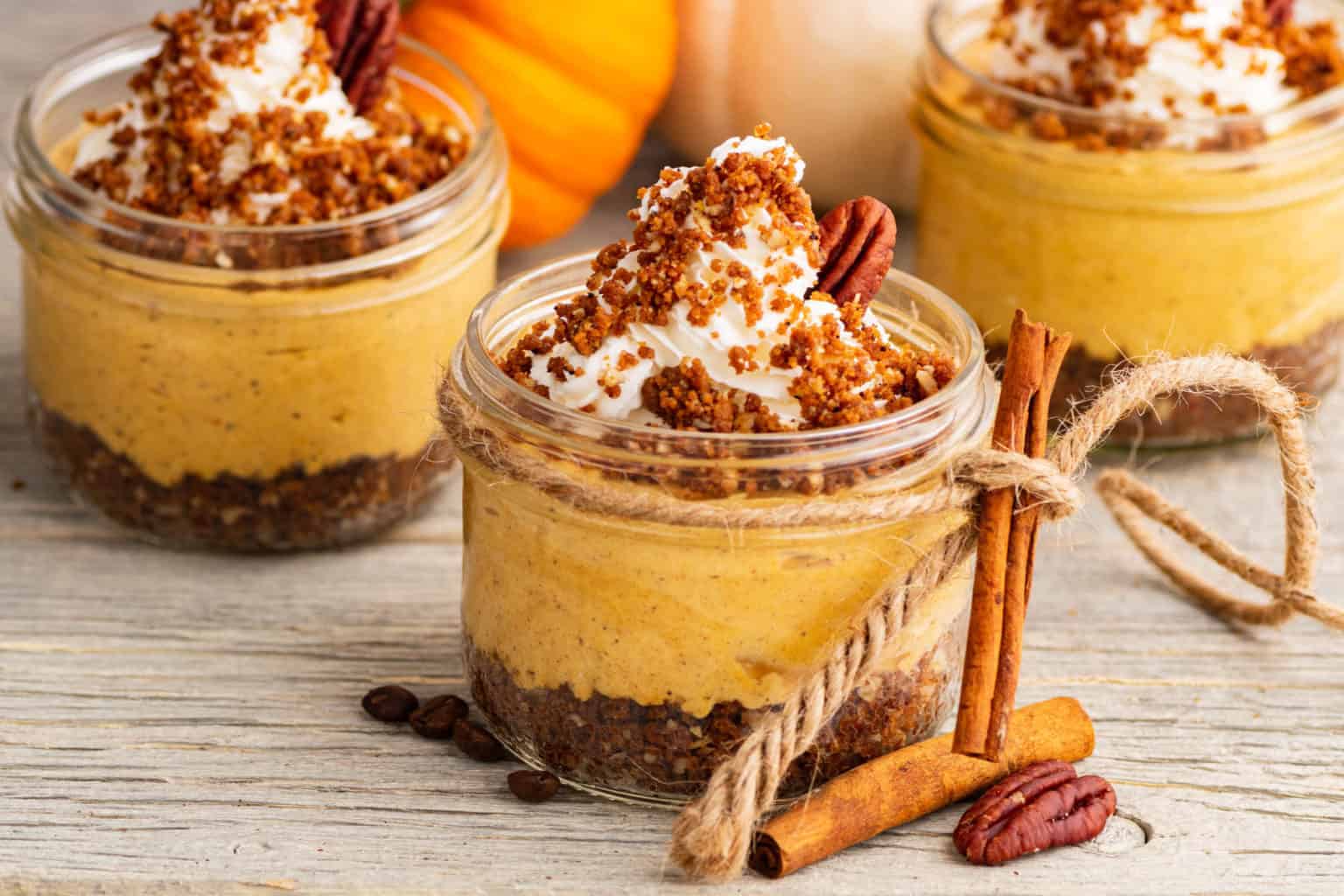 Three jars of pumpkin mousse with whipped cream and crumbled topping are displayed on a wooden surface. Cinnamon sticks are tied together with twine and scattered coffee beans add autumnal touches.