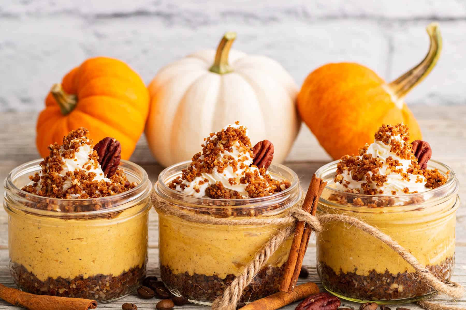 Three individually sized Pumpkin Delight desserts topped with gingersnap crumbs and pecans sit amongst decorative pumpkins.