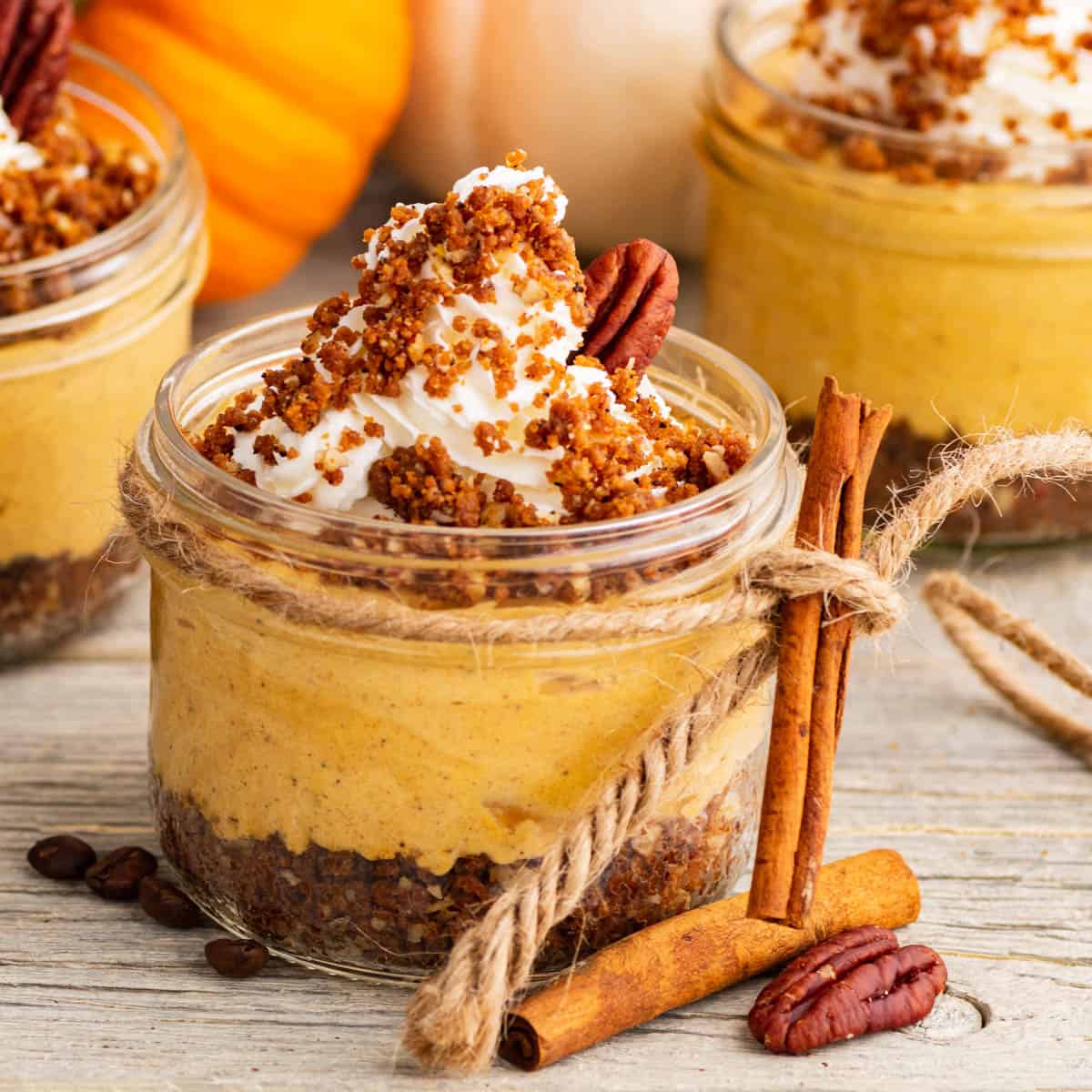 Three individually sized Pumpkin Delight desserts topped with gingersnap crumbs and pecans sit amongst decorative pumpkins.