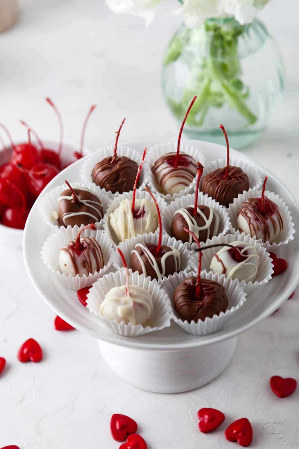 Chocolate cherries on a cake stand in paper cups with red hearts scattered below. 