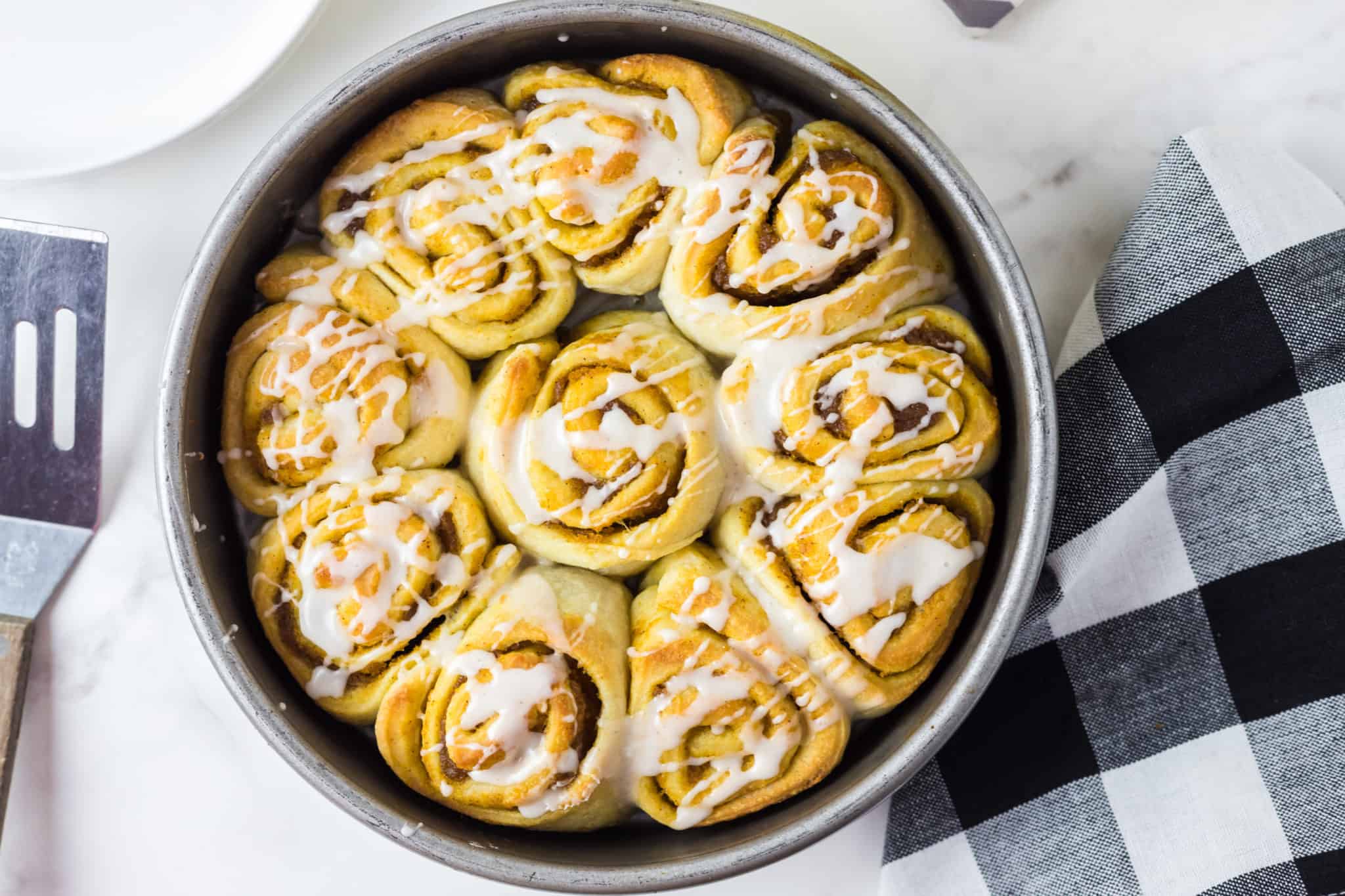 A round baking pan filled with freshly baked cinnamon rolls drizzled with white icing. The cinnamon rolls are golden brown and arranged closely together. A spatula and a black and white checkered cloth are visible beside the pan on a marble surface.