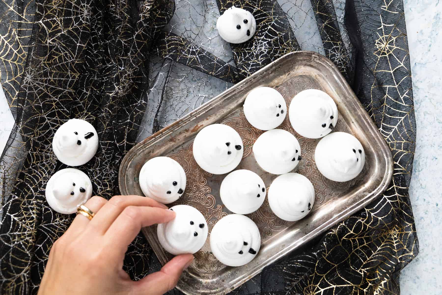 Overhead shot of Meringue Ghosts on a tray, with a hand going to pick one up. 