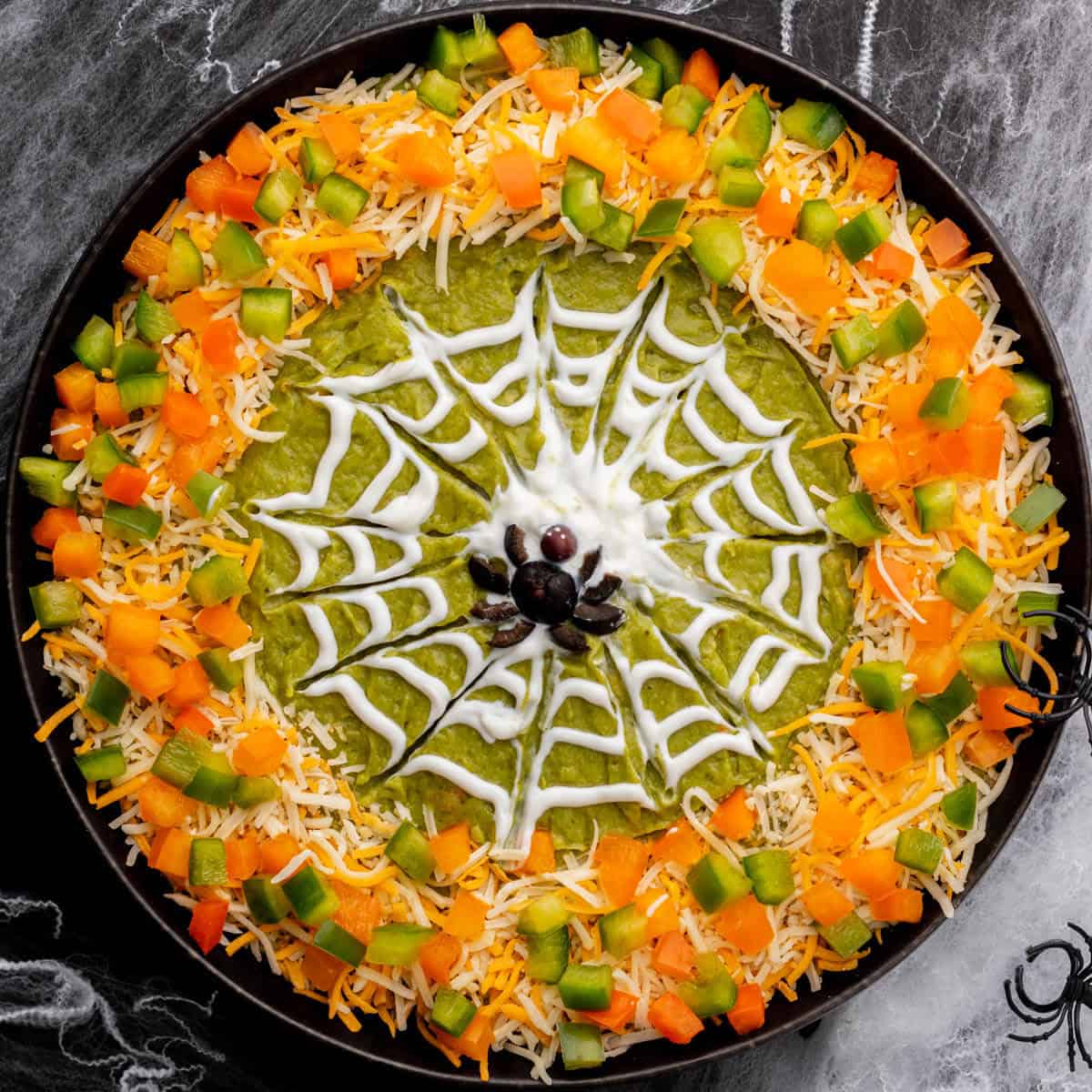 A black bowl containing layered Spider Web Dip decorated with shredded cheese, bell peppers and a sour cream spider web and spiders.
