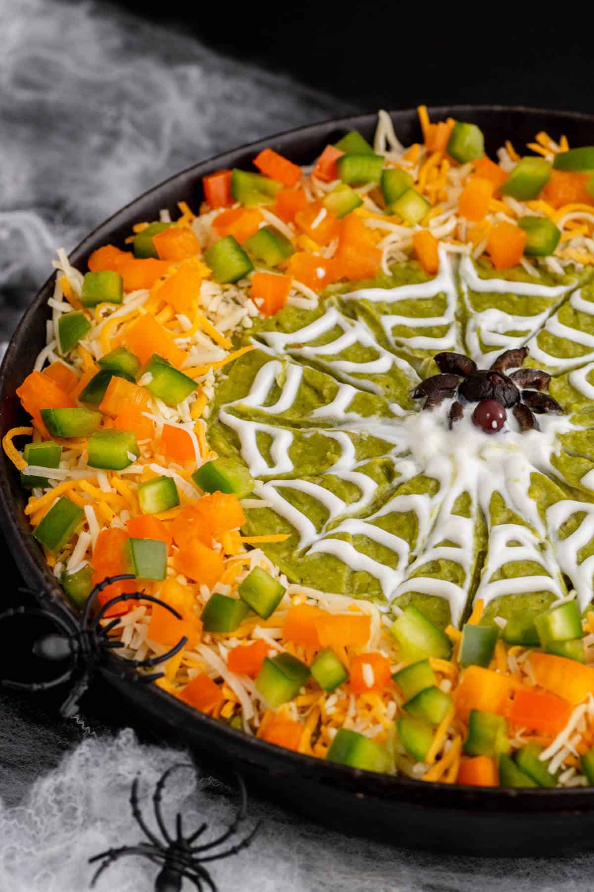 A black bowl containing layered Spider Web Dip decorated with shredded cheese, bell peppers and a sour cream spider web and spiders.