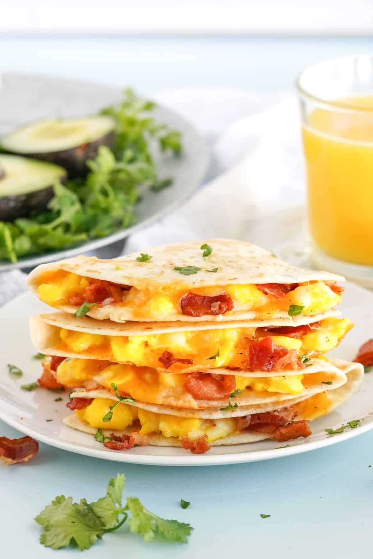 Easy Breakfast Quesadillas stacked on a plate with avocado halved in the background and orange juice. 