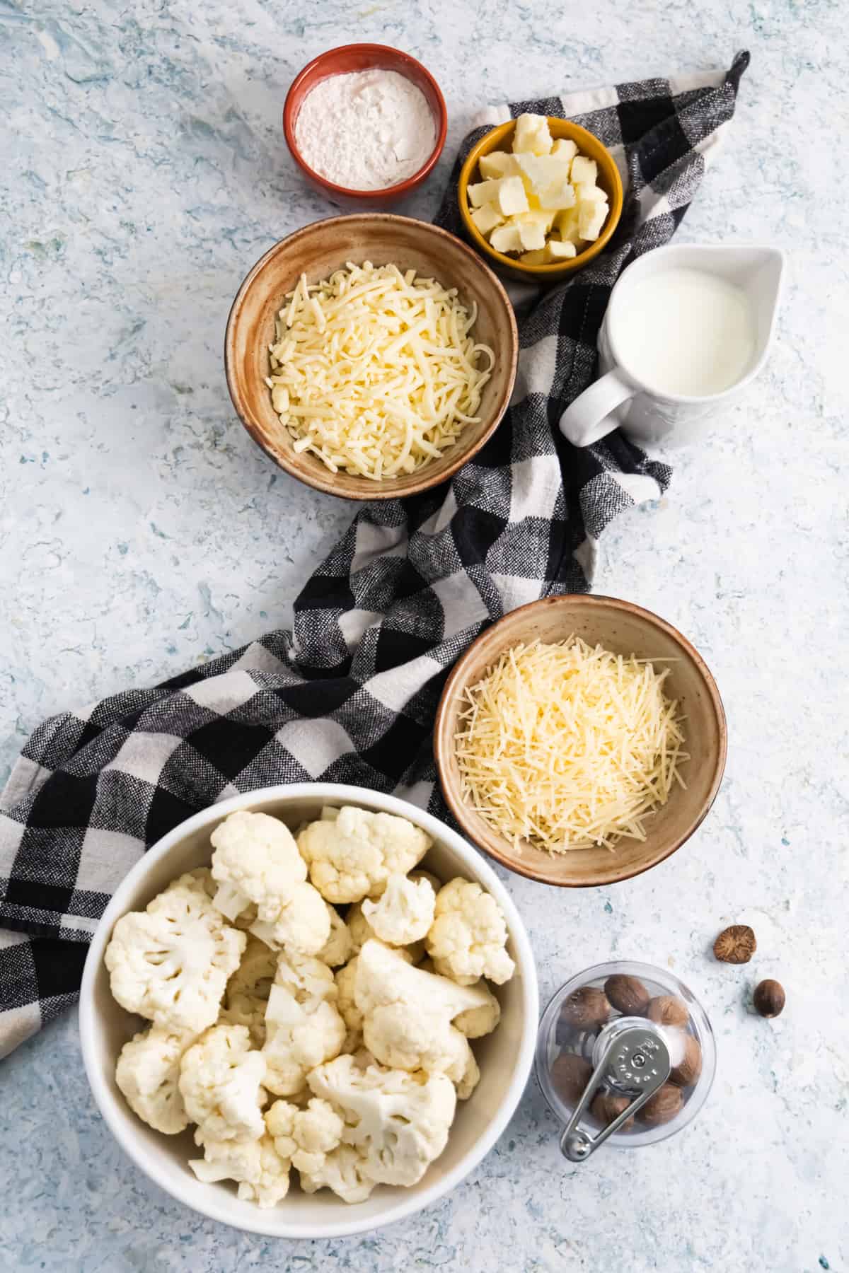 Top view of ingredients, 2 small ramekins with cubed butter and flour, 2 bowls with grated cheese, a small pitcher of milk and a large bowl with cauliflower florets