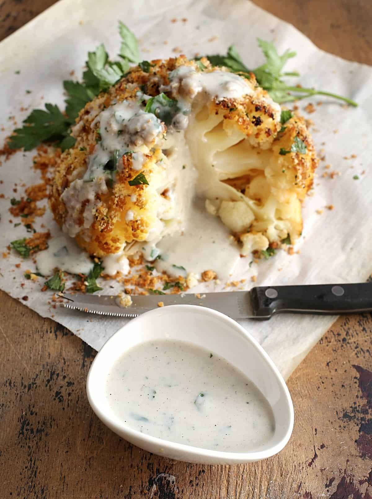 Showing a cut roasted cauliflower with a bowl of Parmesan Cheese sauce and a knife.