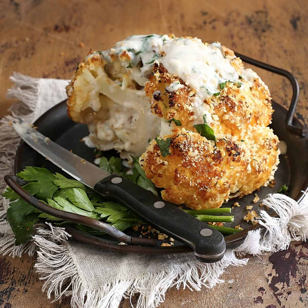 Square photo of a partially cut roasted cauliflower on a plate with a knife