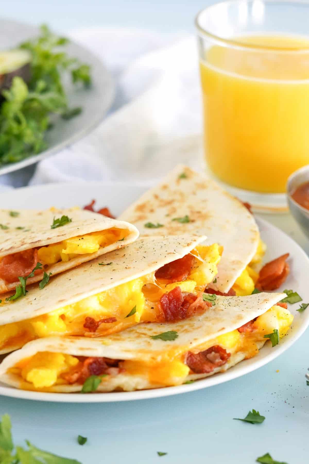 Great looking breakfast quesadillas with bacon, eggs and cheese on a white plate with orange juice in the background. 