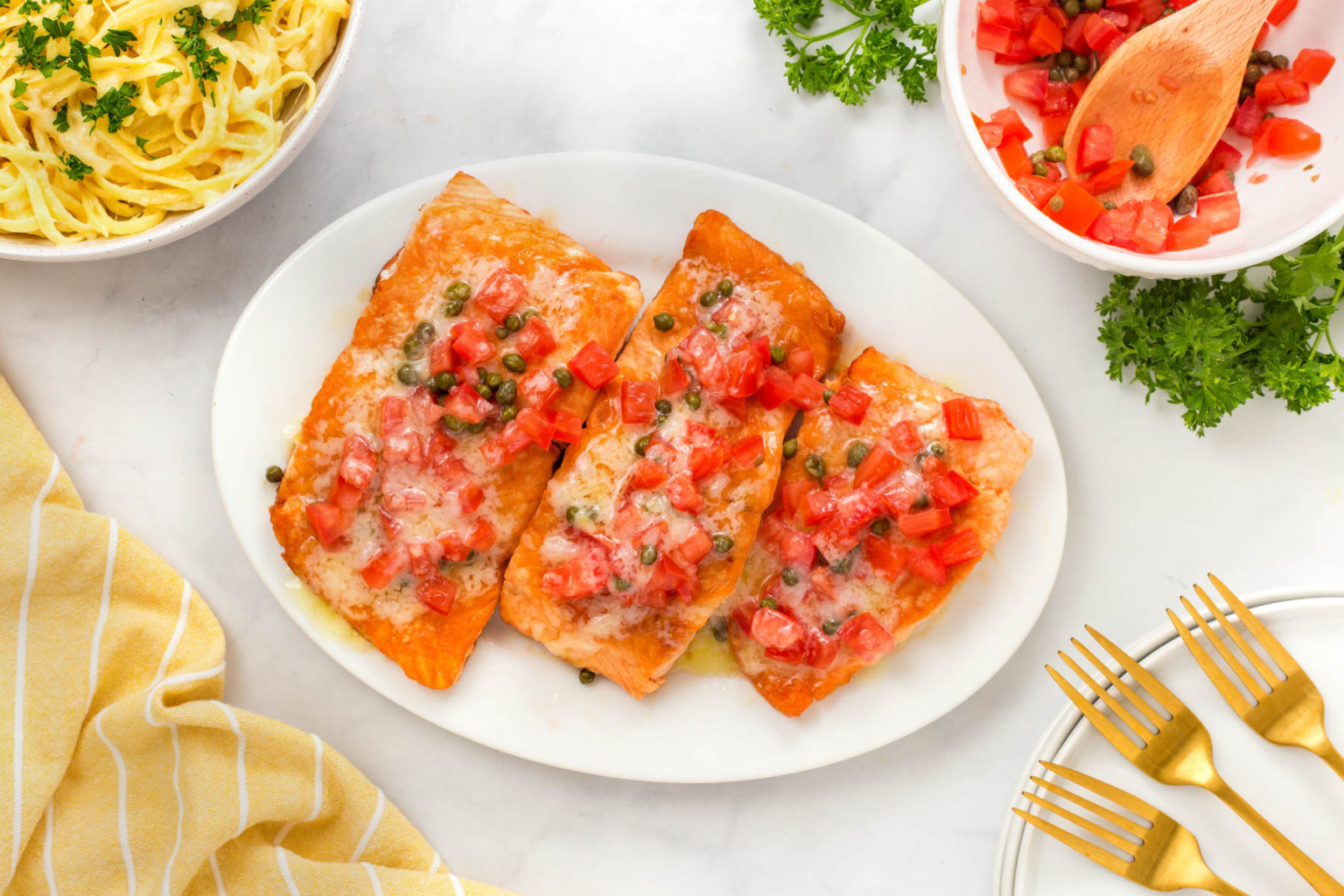 A white oval plate with three pieces of salmon topped with diced tomatoes, capers, and melted cheese. Surrounding the plate are a bowl of pasta, a dish with tomato mix, a bunch of parsley, golden forks, and a yellow striped cloth on a marble surface.