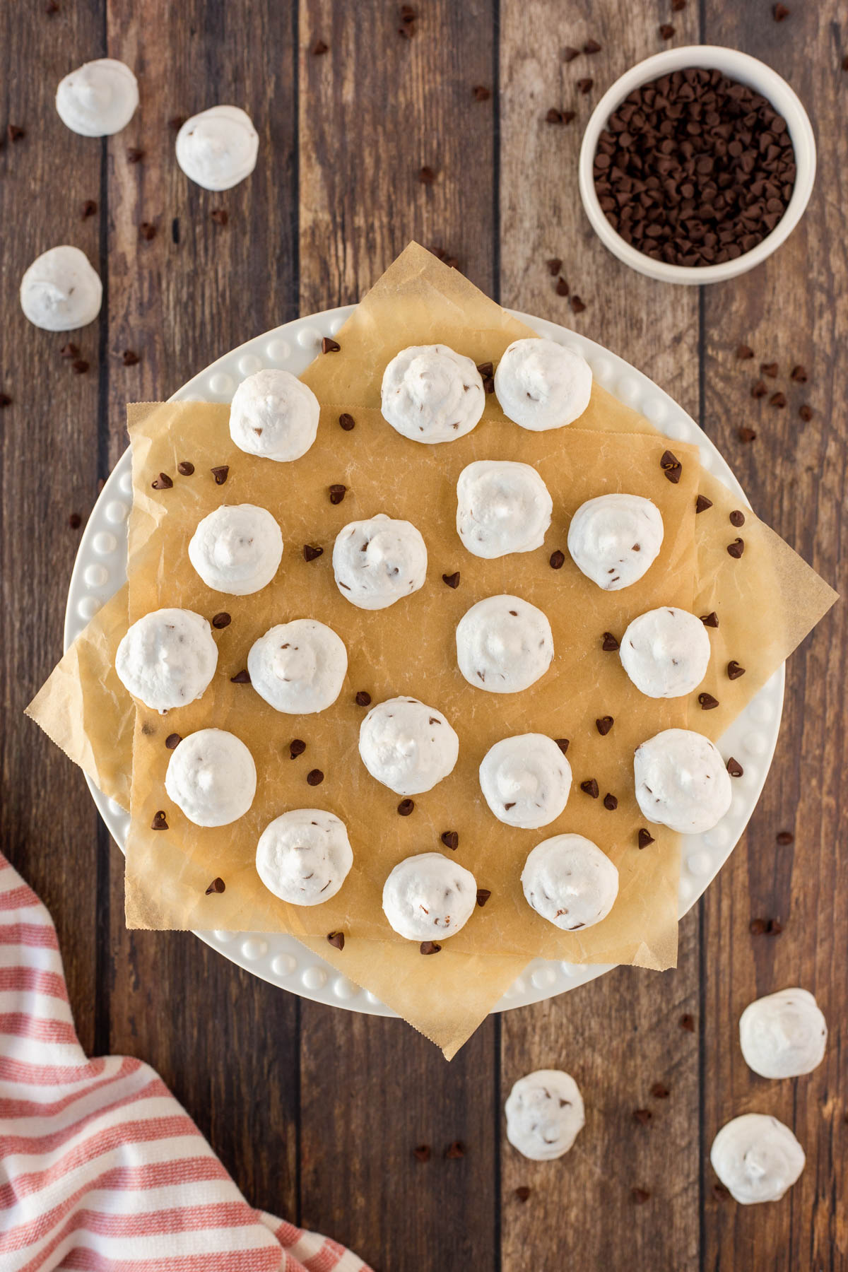 Mini chocolate Chip Meringue Cookies on a white cake platter with mini chocolate chips around the cookies.