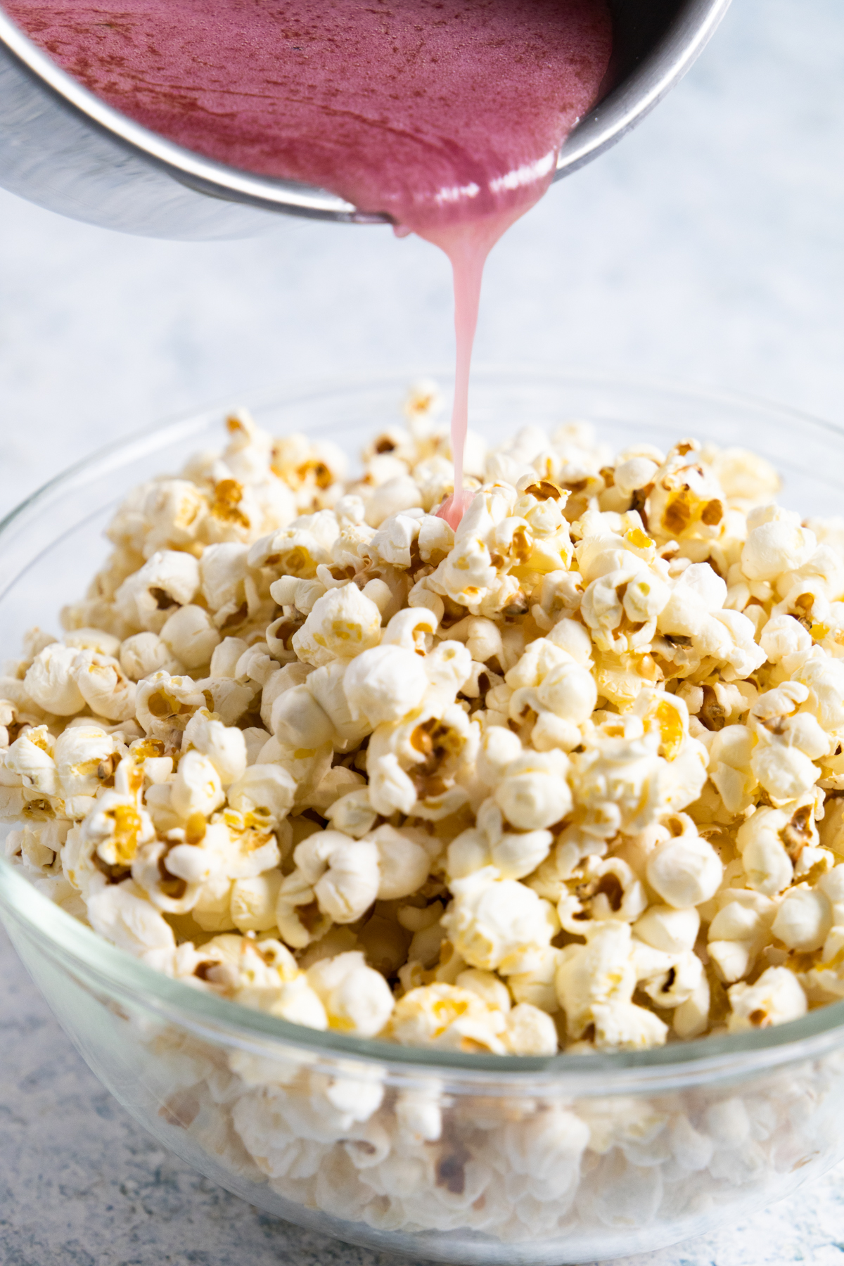 side view of plain popcorn in a transparent bowl with red syrup being poured. 