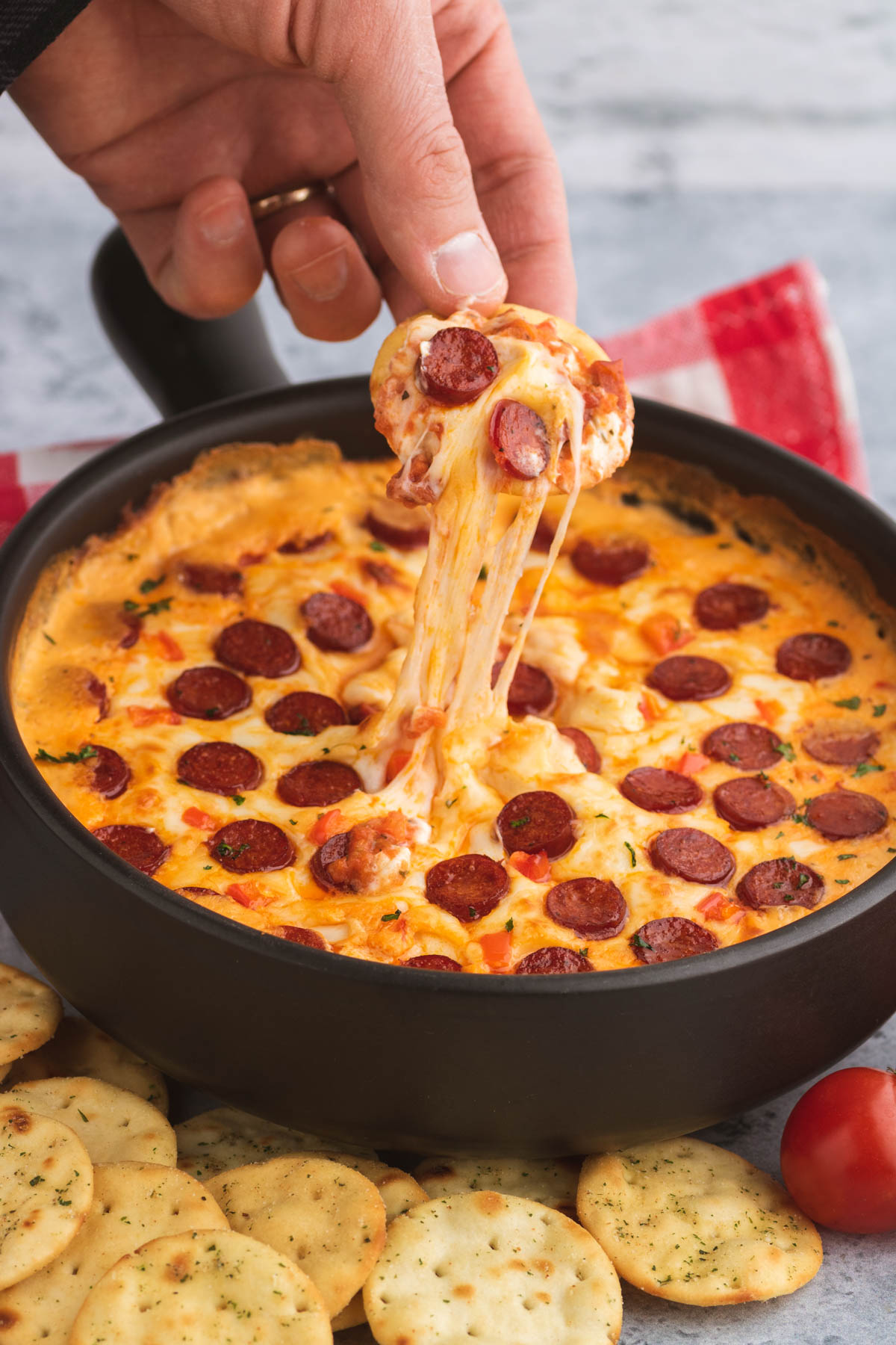 A hand dips a cracker into hot bubbly pizza dip, leaving a trail of melted cheese.