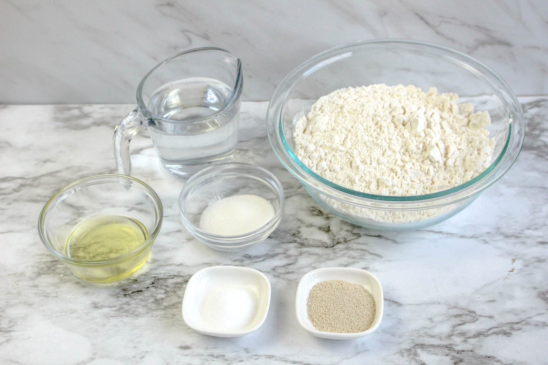 All the ingredients to make bread machine pizza dough in glass bowls on a marble counter.
