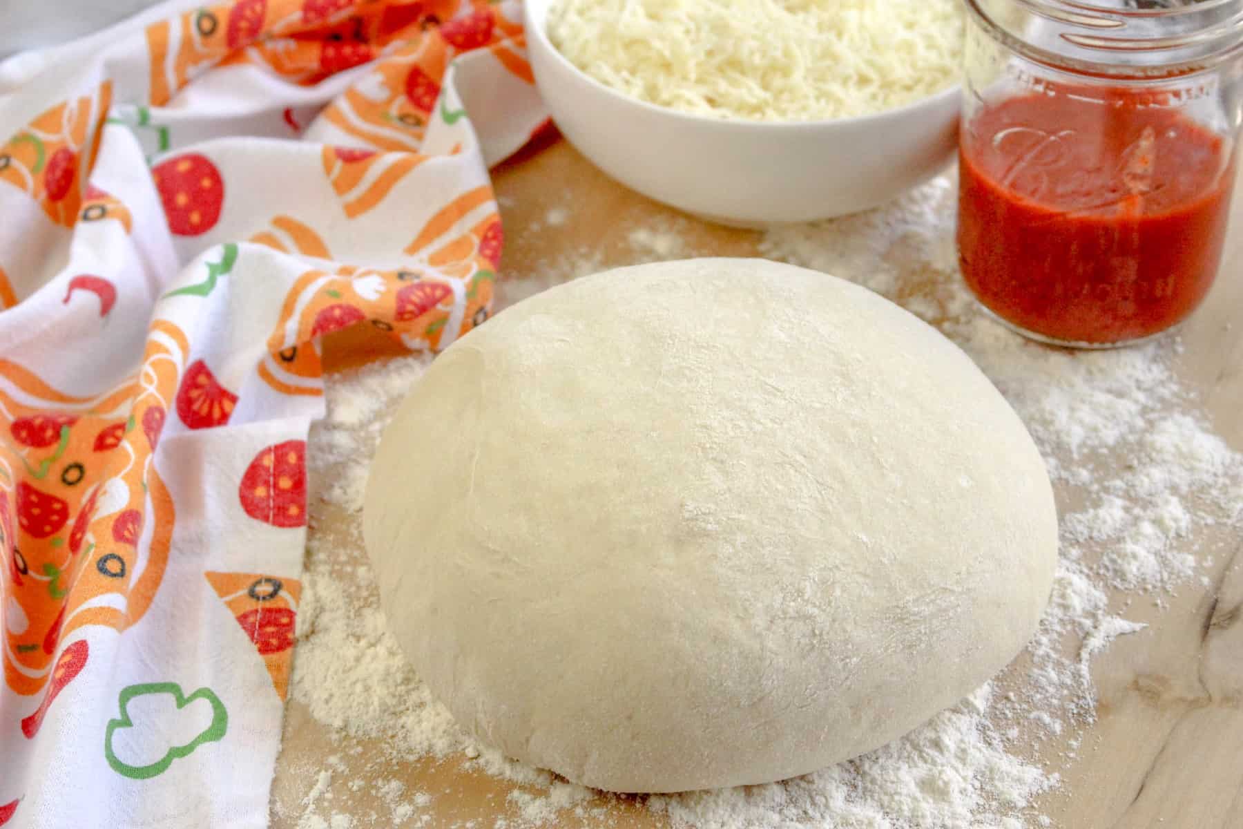 Horizontal picture of pizza dough with a pizza tea towel, cheese and tomato sauce on the side. 