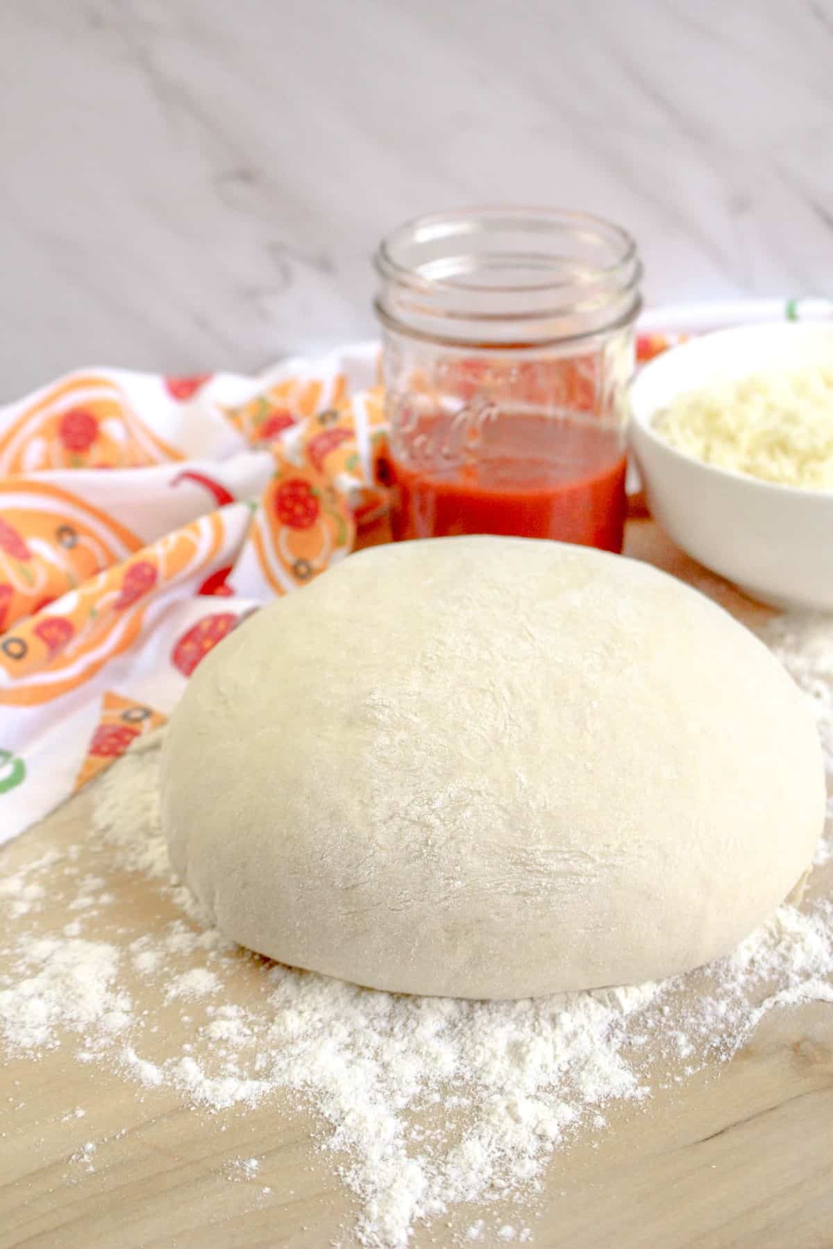 Ball of pizza dough resting on a floured surface with pizza sauce and grated cheese in bowls behind the dough.